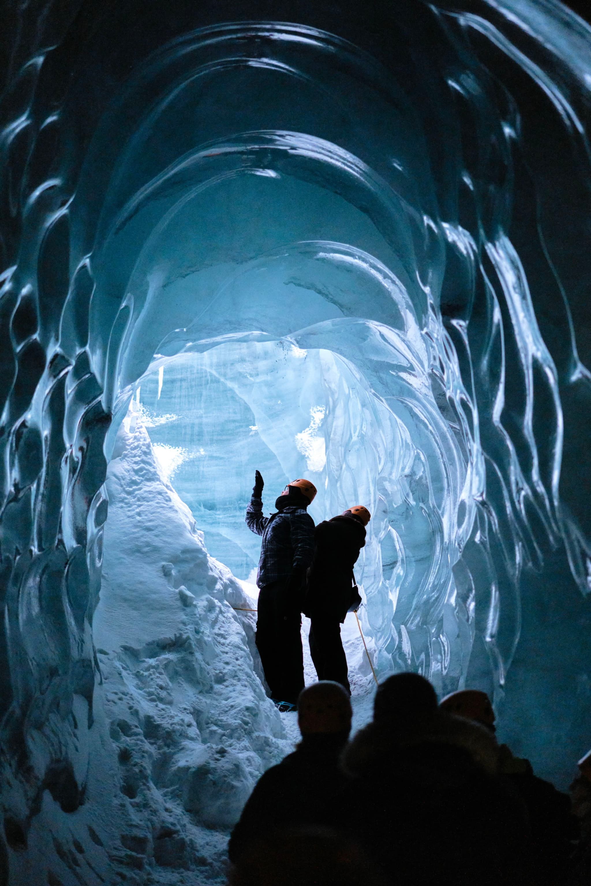 People exploring an ice cave with textured, blue walls and a snowy floor, illuminated by natural light