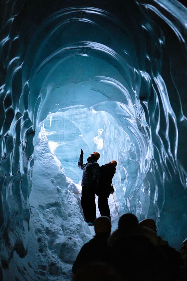 People exploring an ice cave with textured, blue walls and a snowy floor, illuminated by natural light