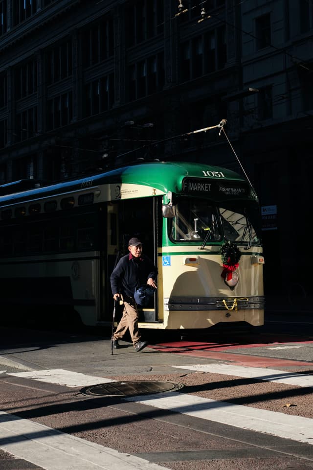 A man walking across a street in front of a vintage green and cream streetcar, with buildings in the background