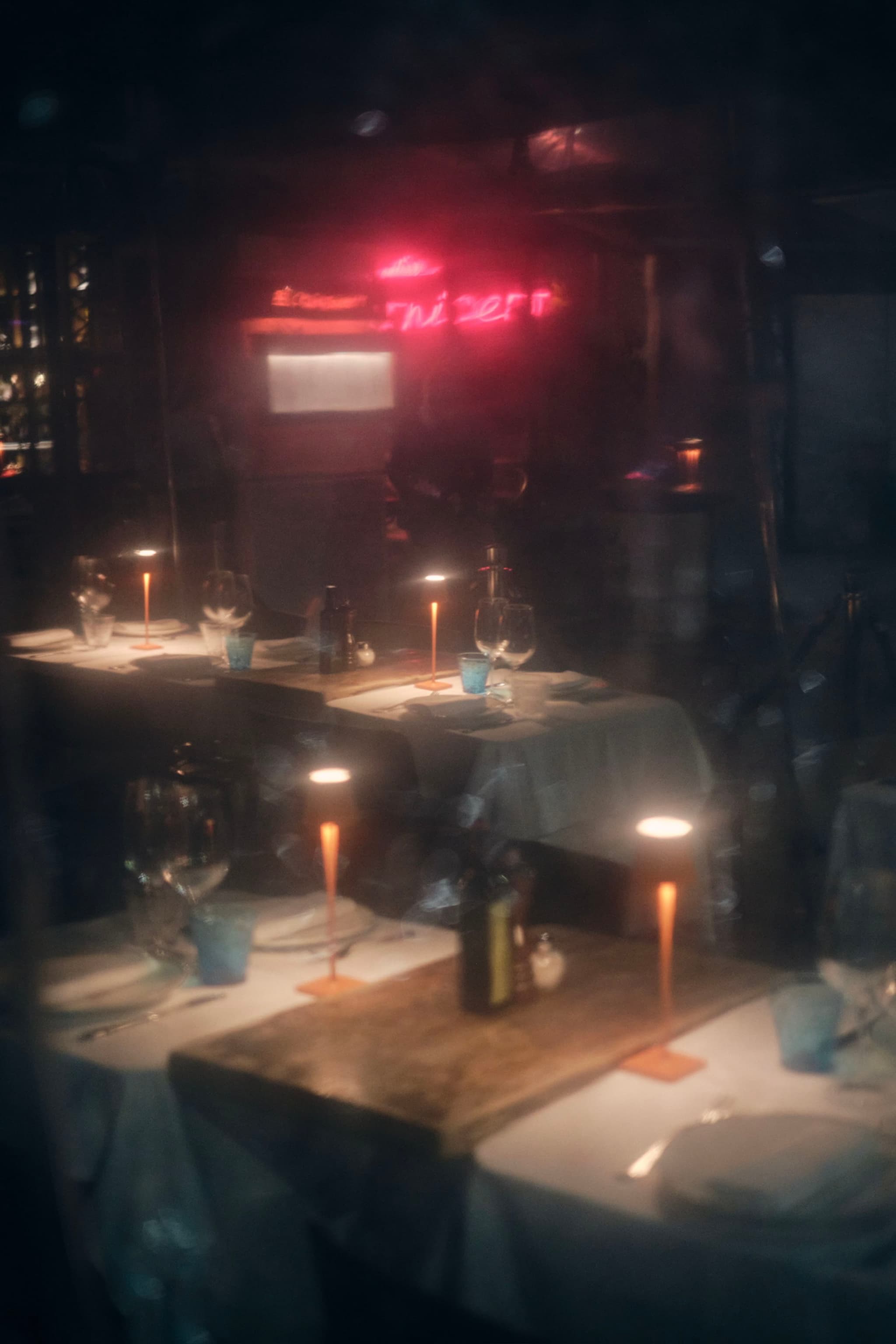 A dimly lit restaurant interior with tables set for dining, featuring candles and wine glasses, and a neon sign in the background