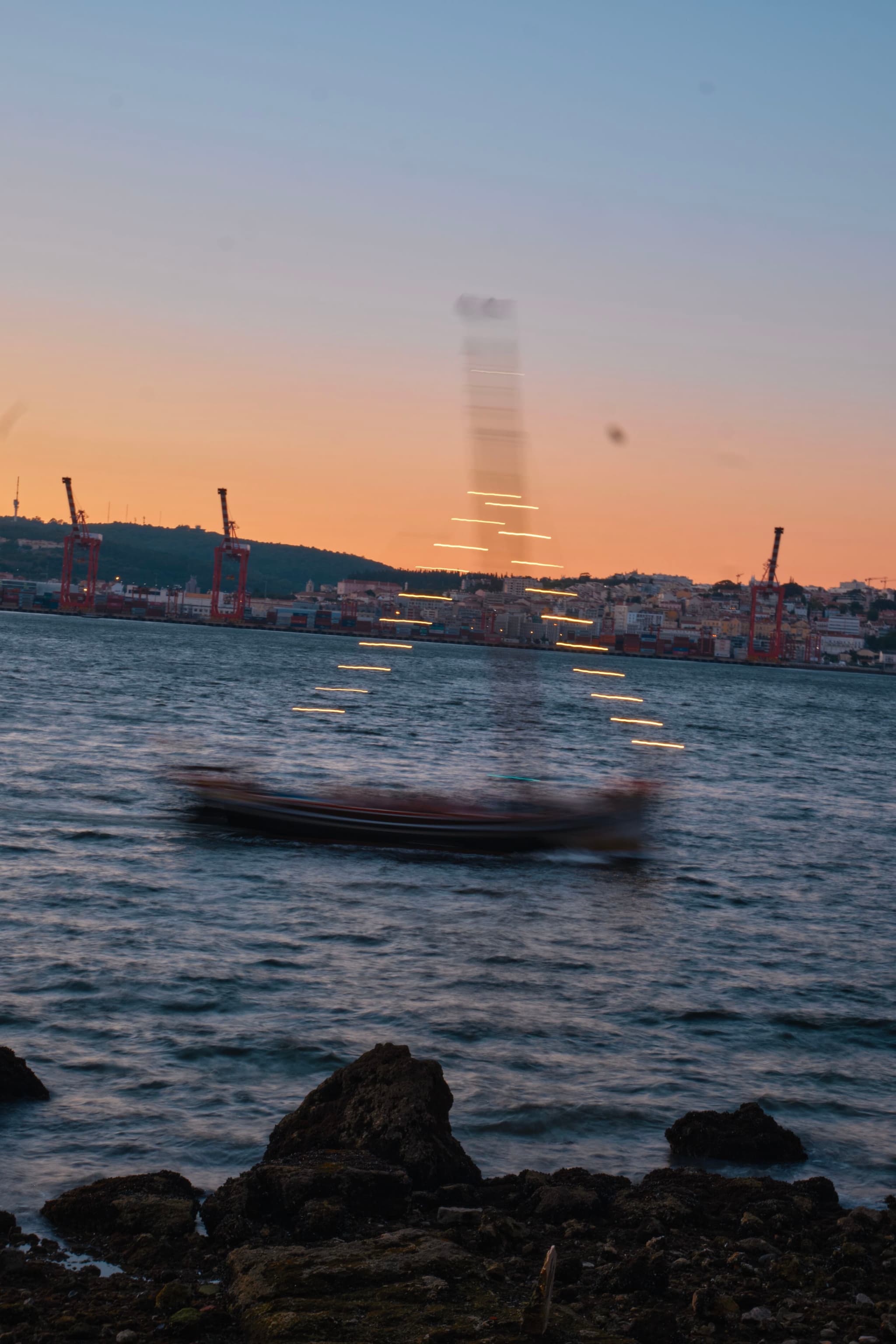 A blurred boat with lights moves across the water at sunset, with a city and cranes in the background