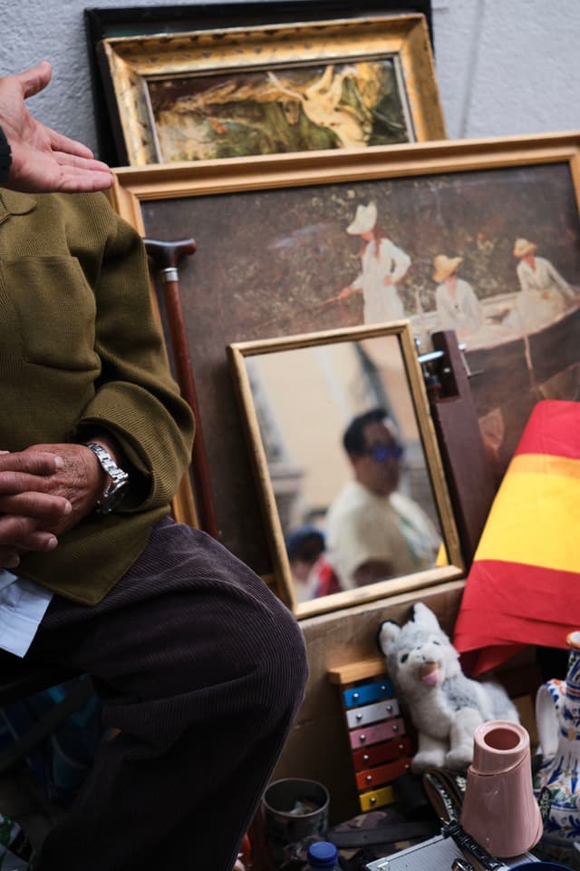 A collection of framed paintings and a mirror reflecting a person, with a seated individual partially visible. A small ceramic cat and a stack of colorful boxes are in the foreground, alongside a red and yellow striped cloth