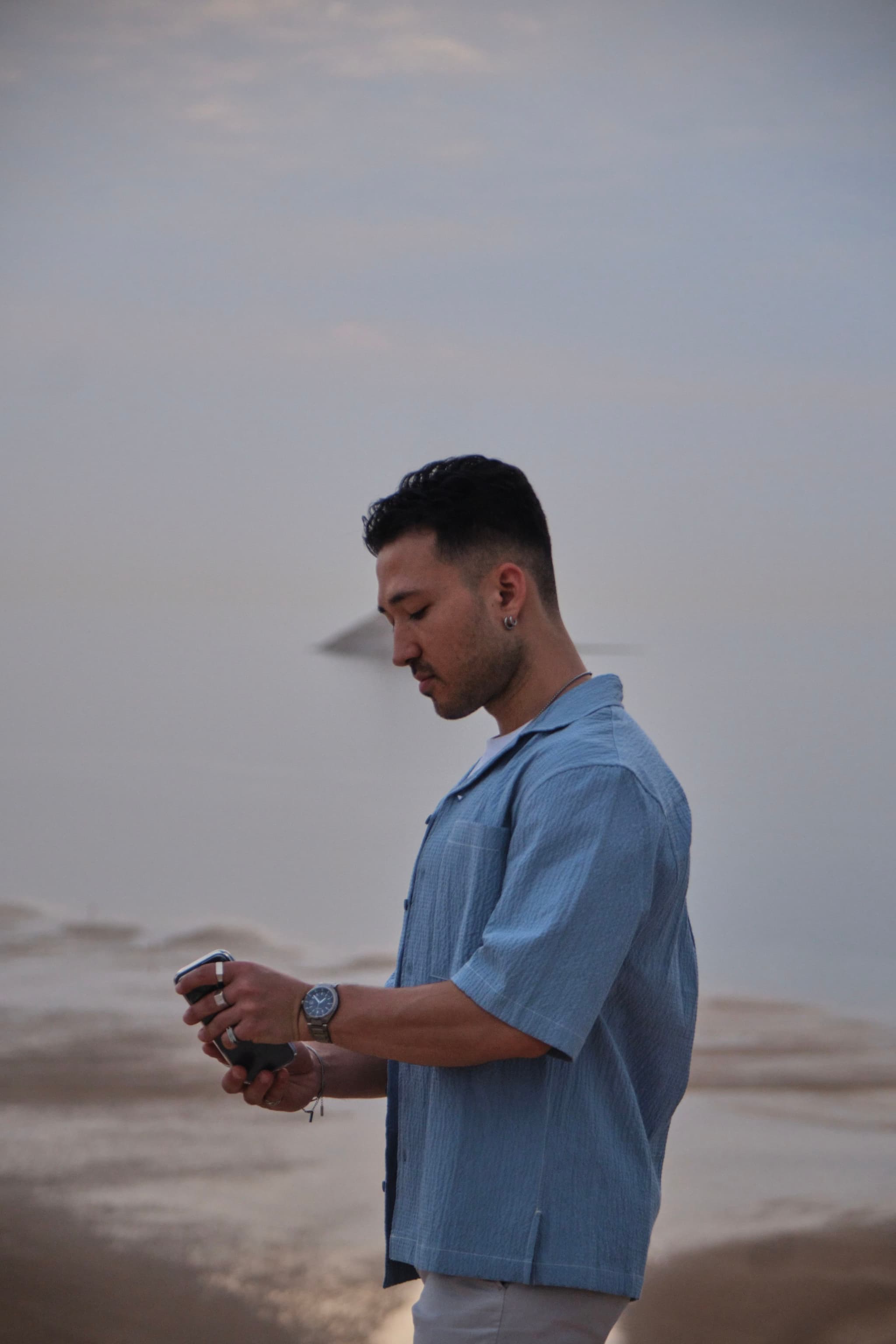 A person in a blue shirt stands on a beach, holding a camera, with a calm sea and cloudy sky in the background