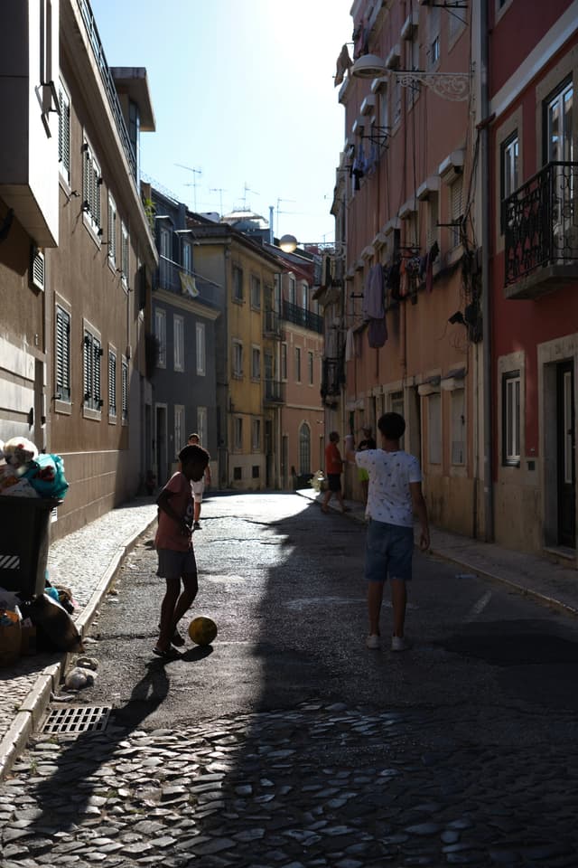A narrow, sunlit street with colorful buildings on either side, where two children are playing with a soccer ball