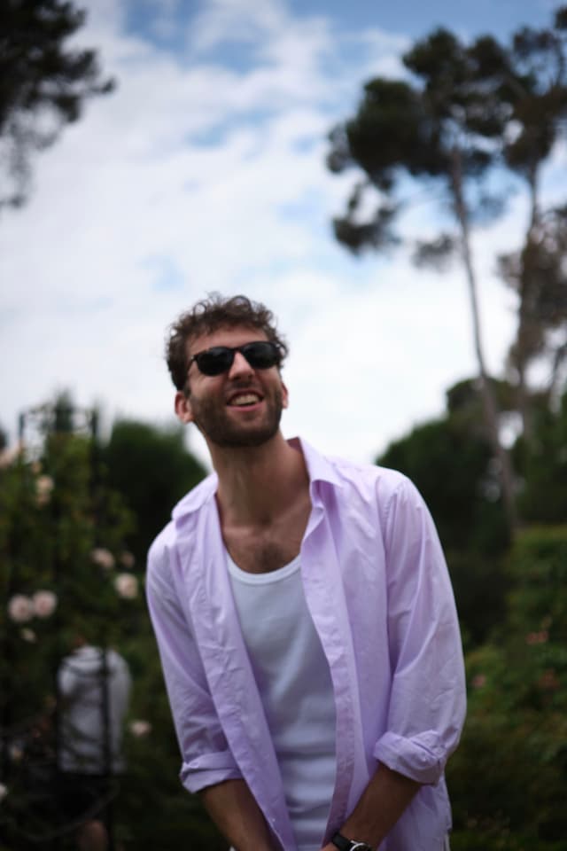A person wearing sunglasses and a light purple shirt stands outdoors, smiling, with trees and a cloudy sky in the background