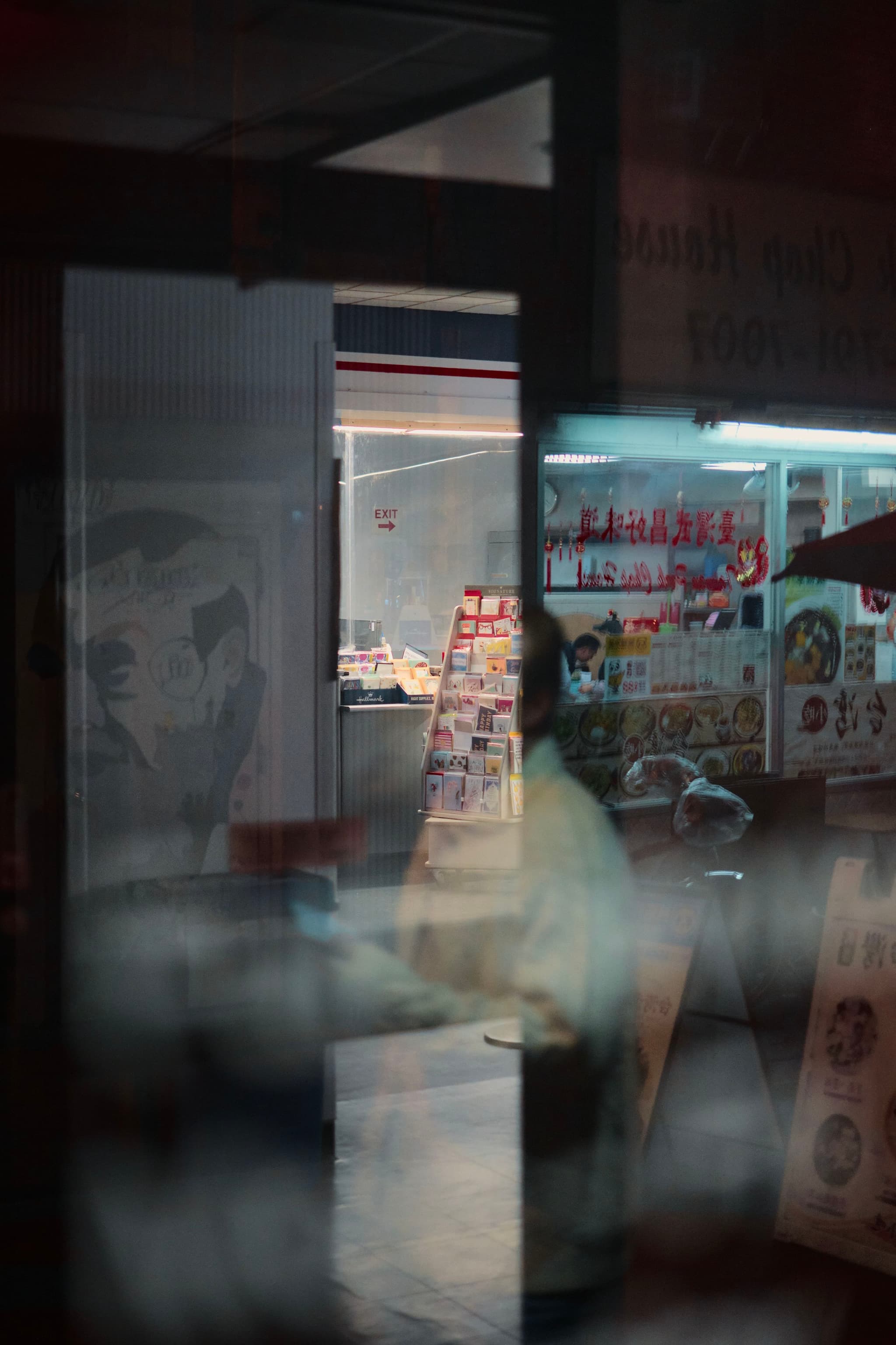 A dimly lit interior of a restaurant with a menu board and food displays, seen through a reflective glass window