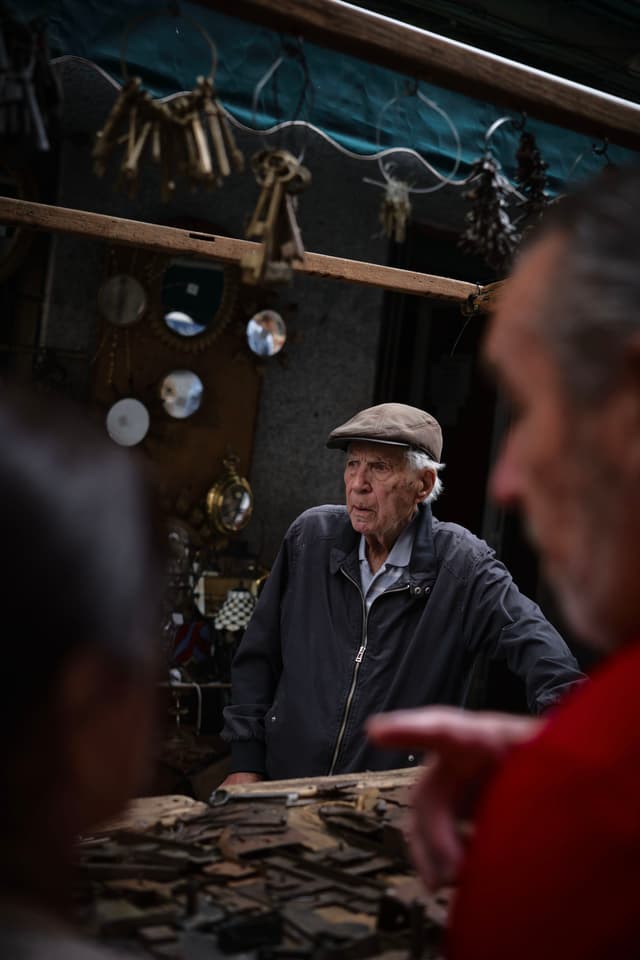 An elderly man wearing a cap stands behind a table with various items, possibly at a market. The background includes hanging objects and mirrors, and there are two people in the foreground, one pointing