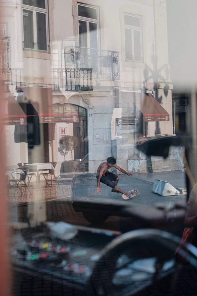 A skateboarder performing a trick in an urban setting, reflected in a window with a view of a street and buildings