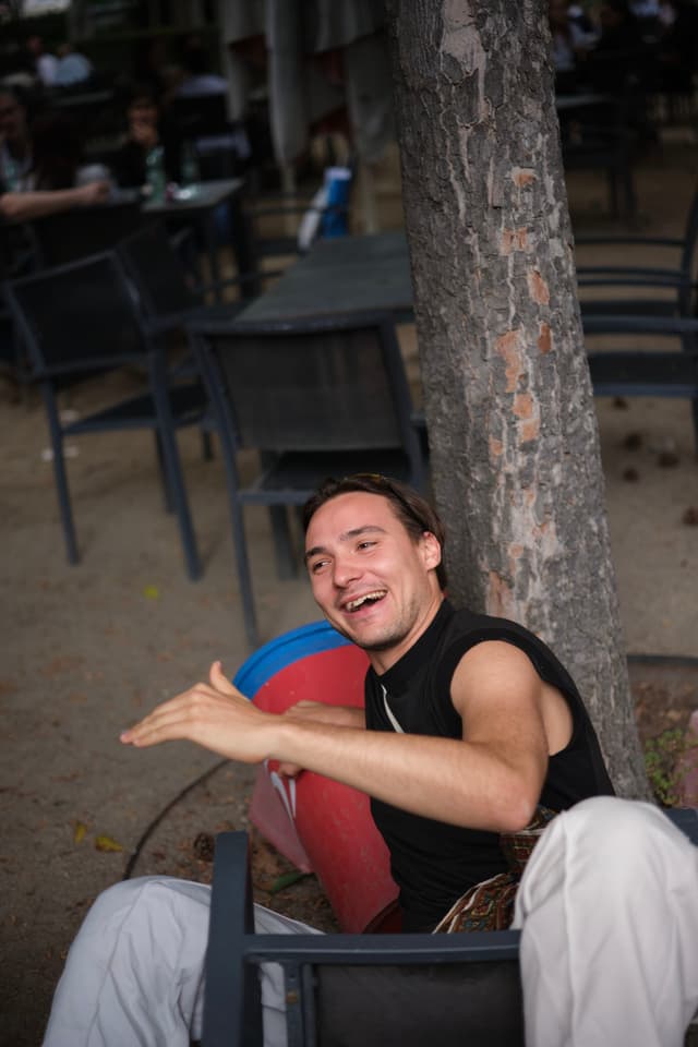 A person sitting outdoors, smiling and gesturing with their hands, surrounded by chairs and a tree