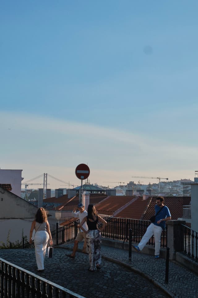 Four people stand on a cobblestone path with a view of a cityscape, including rooftops and a bridge in the background, under a clear blue sky