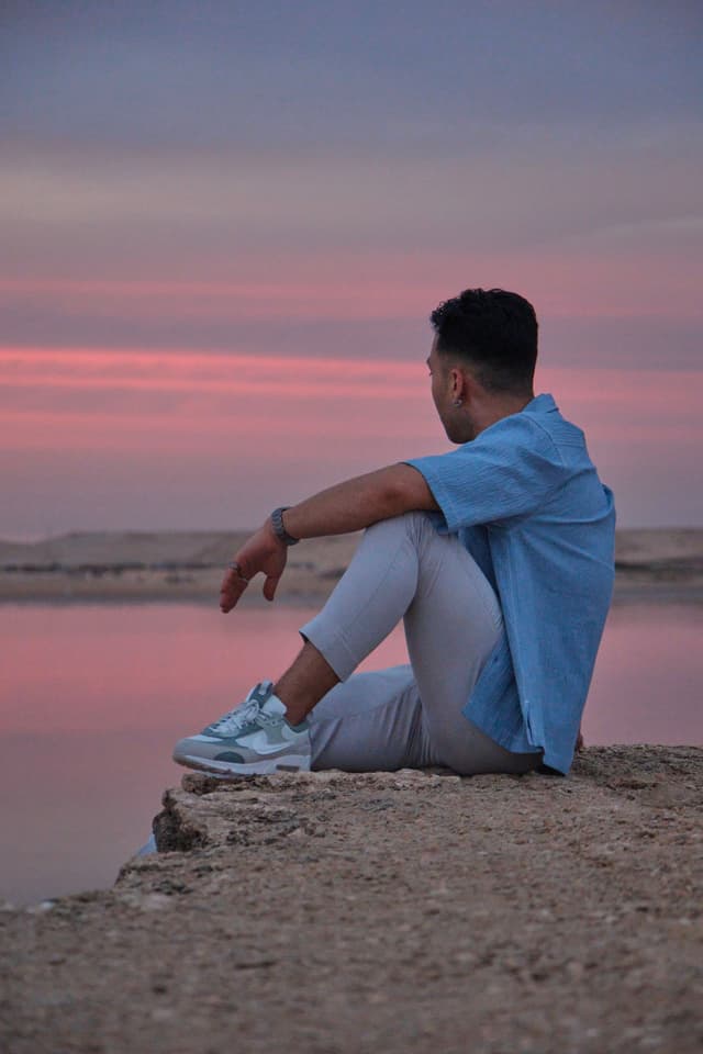 A person in a blue shirt and light pants sits on a rocky surface, gazing at a serene sunset over a calm body of water
