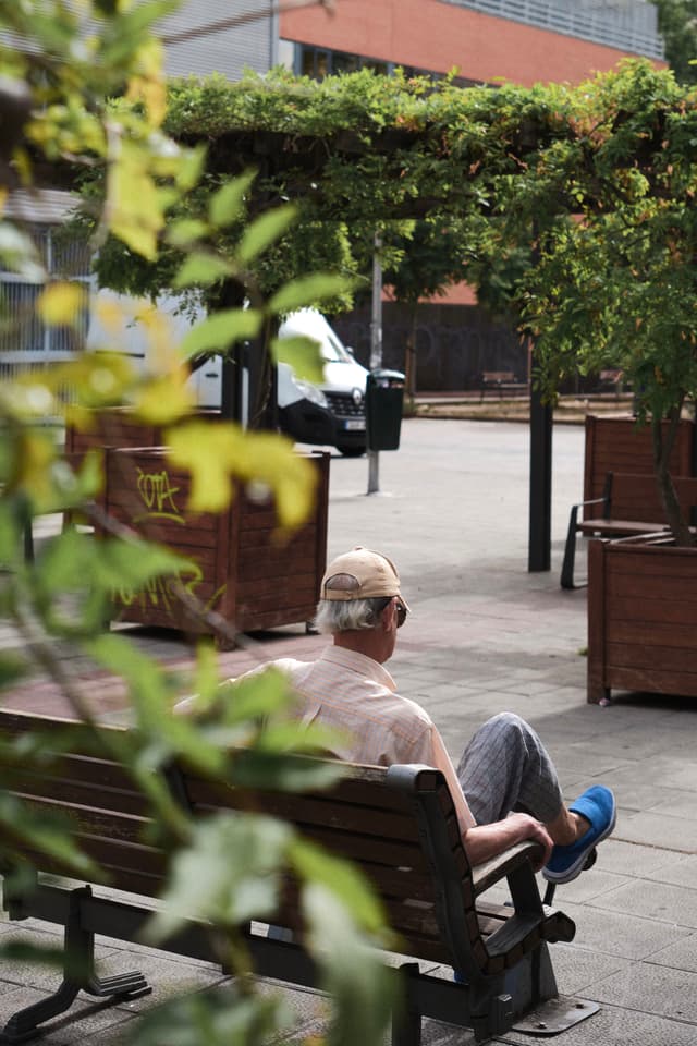 An elderly person wearing a cap sits on a bench in a park-like setting, surrounded by greenery and planters, with a paved area and buildings in the background