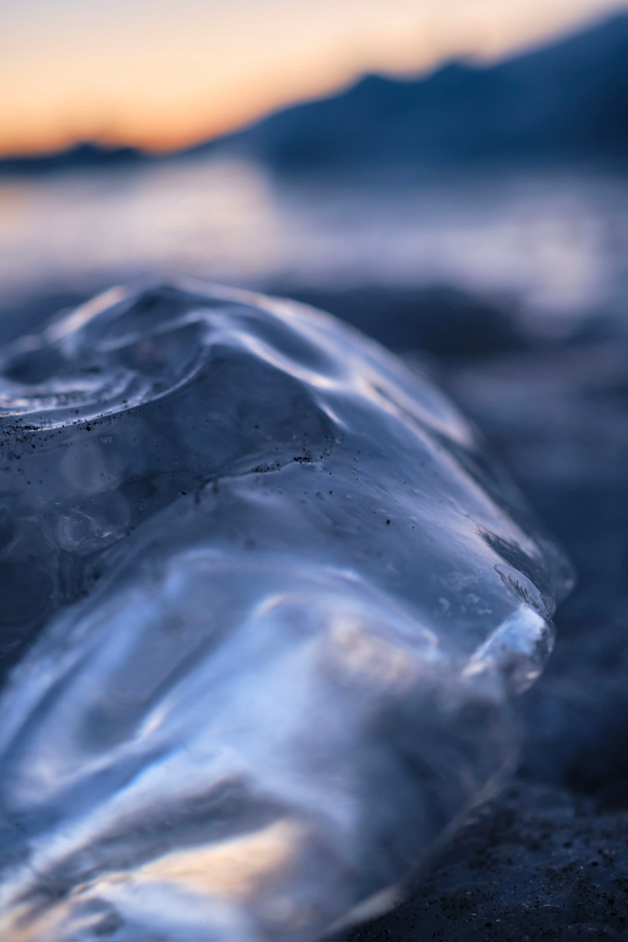 A close-up of a smooth, translucent ice formation on a beach, with a blurred background of water and a sunset sky