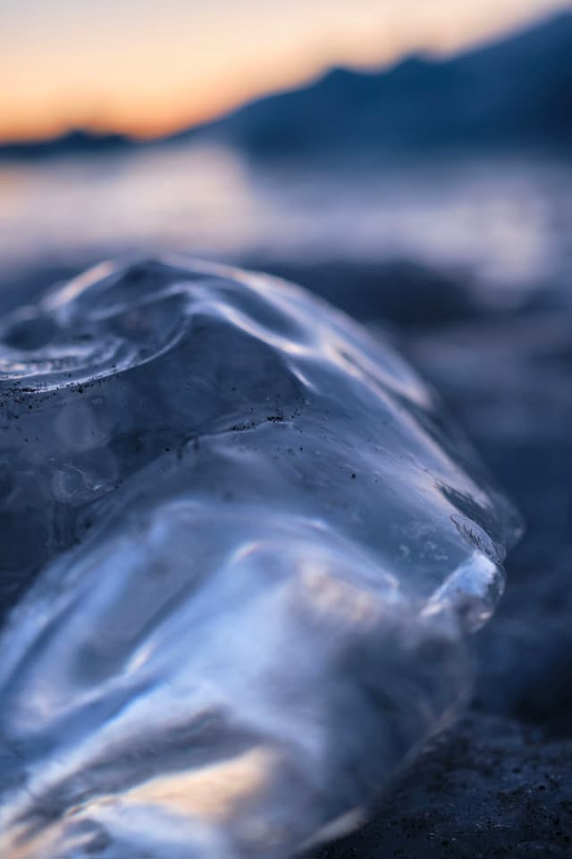 A close-up of a smooth, translucent ice formation on a beach, with a blurred background of water and a sunset sky