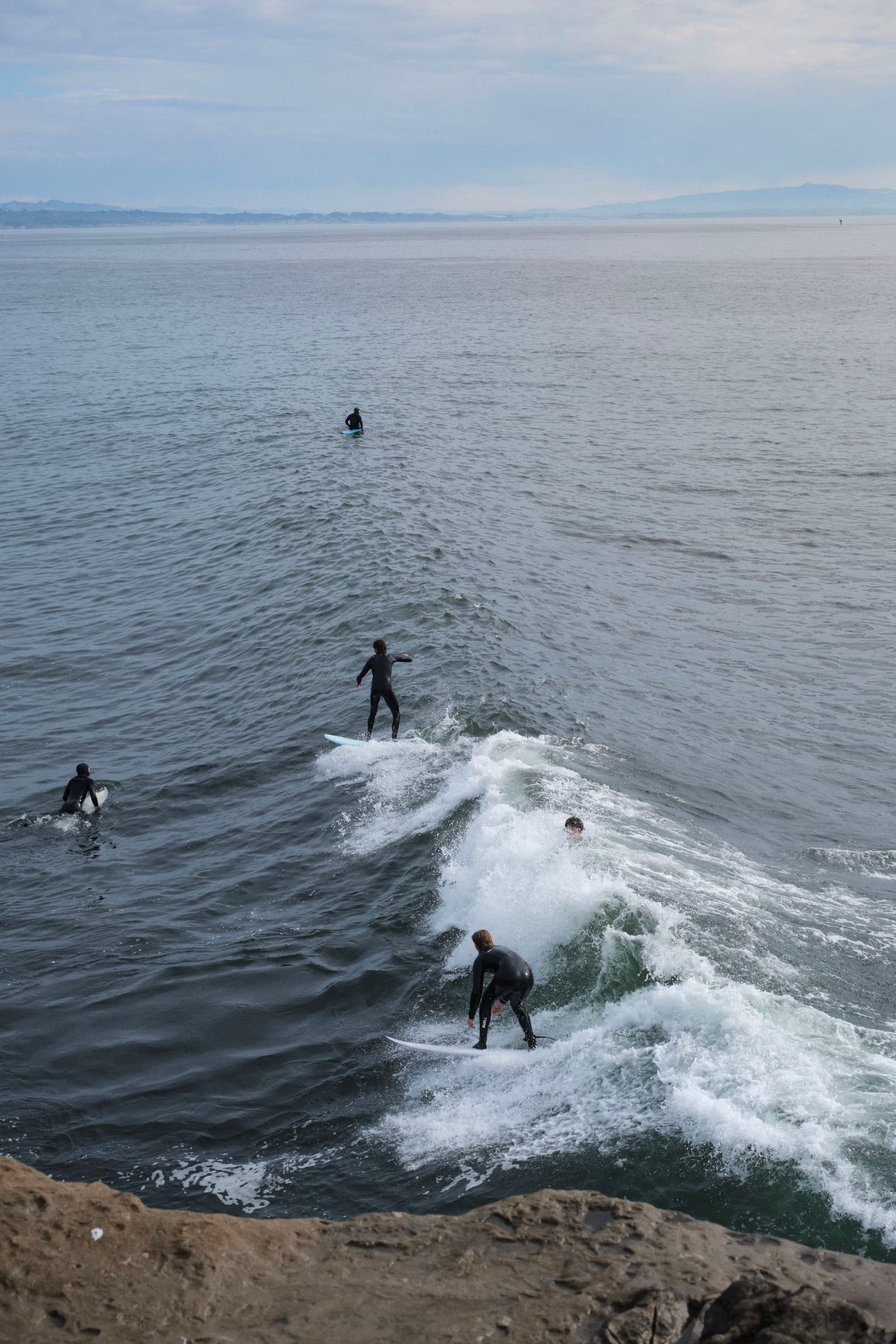 Surfers riding waves near a rocky shoreline with a calm ocean and cloudy sky in the background