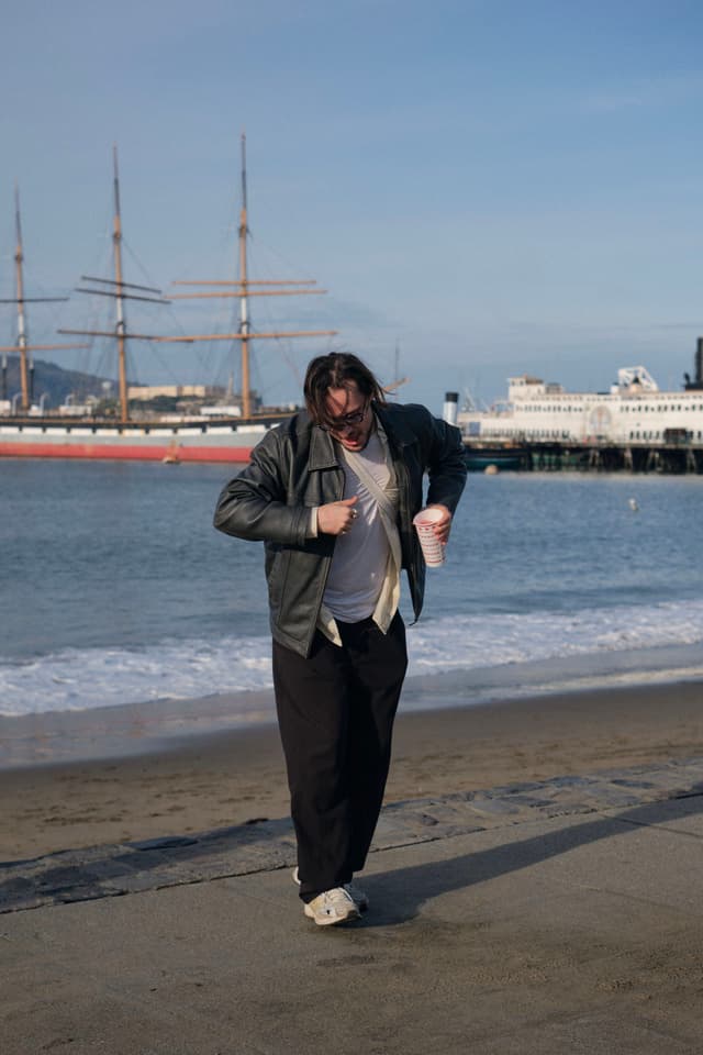 A person walking on a beach near the water, wearing a jacket and holding a cup, with ships and a pier in the background