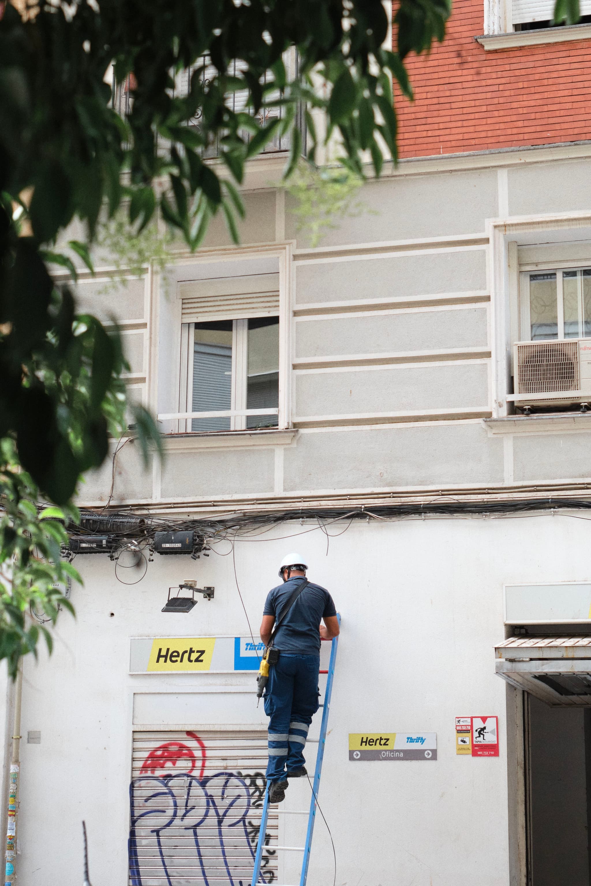 A worker on a ladder is fixing cables on the exterior of a building with a Hertz sign, surrounded by some graffiti and greenery