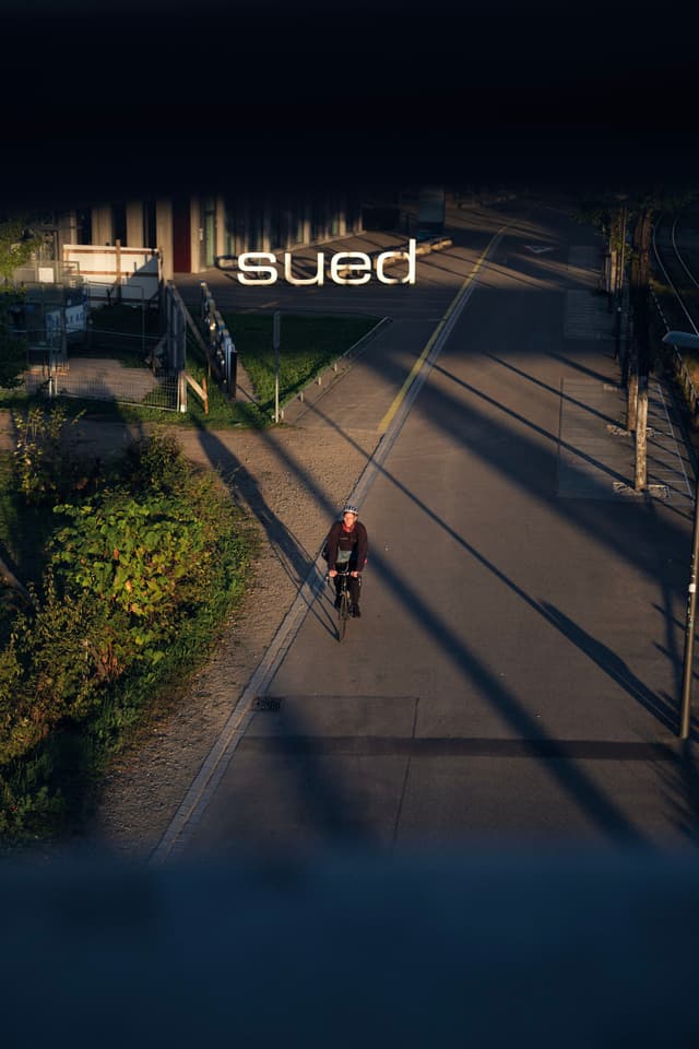 A person riding a bicycle on a sunlit path, with long shadows cast across the ground and the word sued visible above