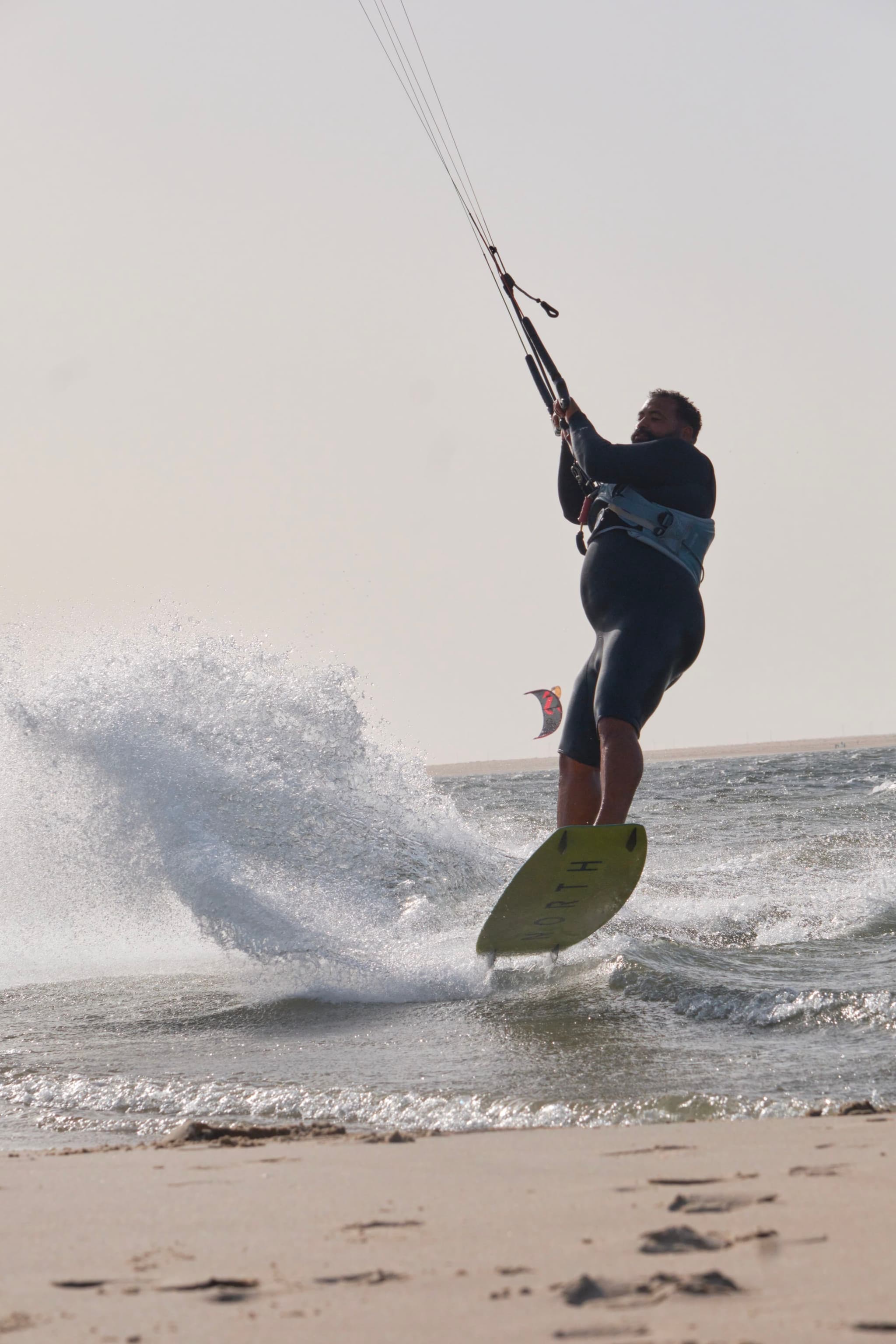 A person is kiteboarding on the water, creating a splash, with a sandy beach in the foreground