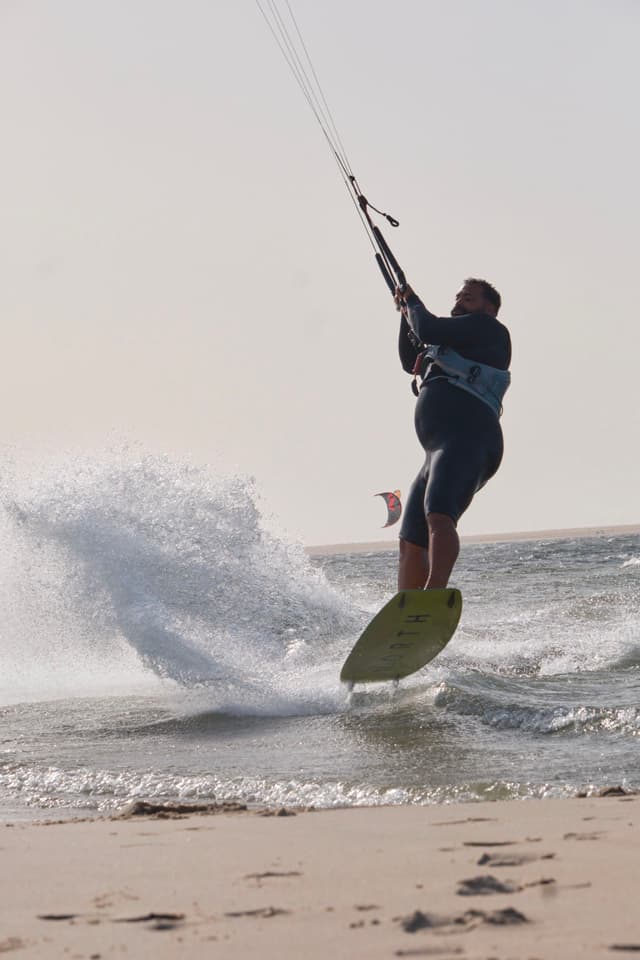 A person is kiteboarding on the water, creating a splash, with a sandy beach in the foreground