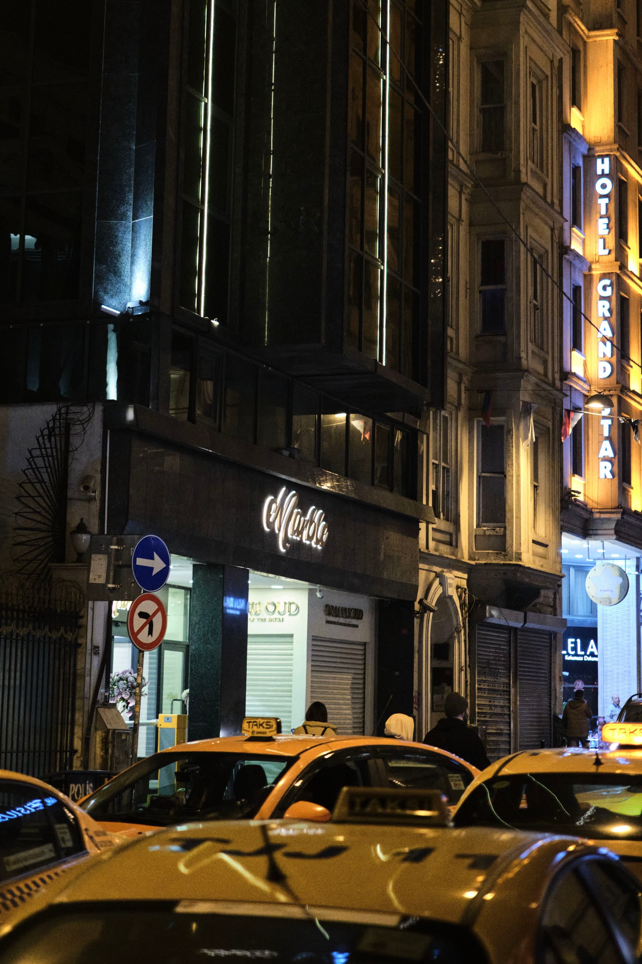 A nighttime street scene with several yellow taxis in the foreground, illuminated buildings, and a hotel sign in the background