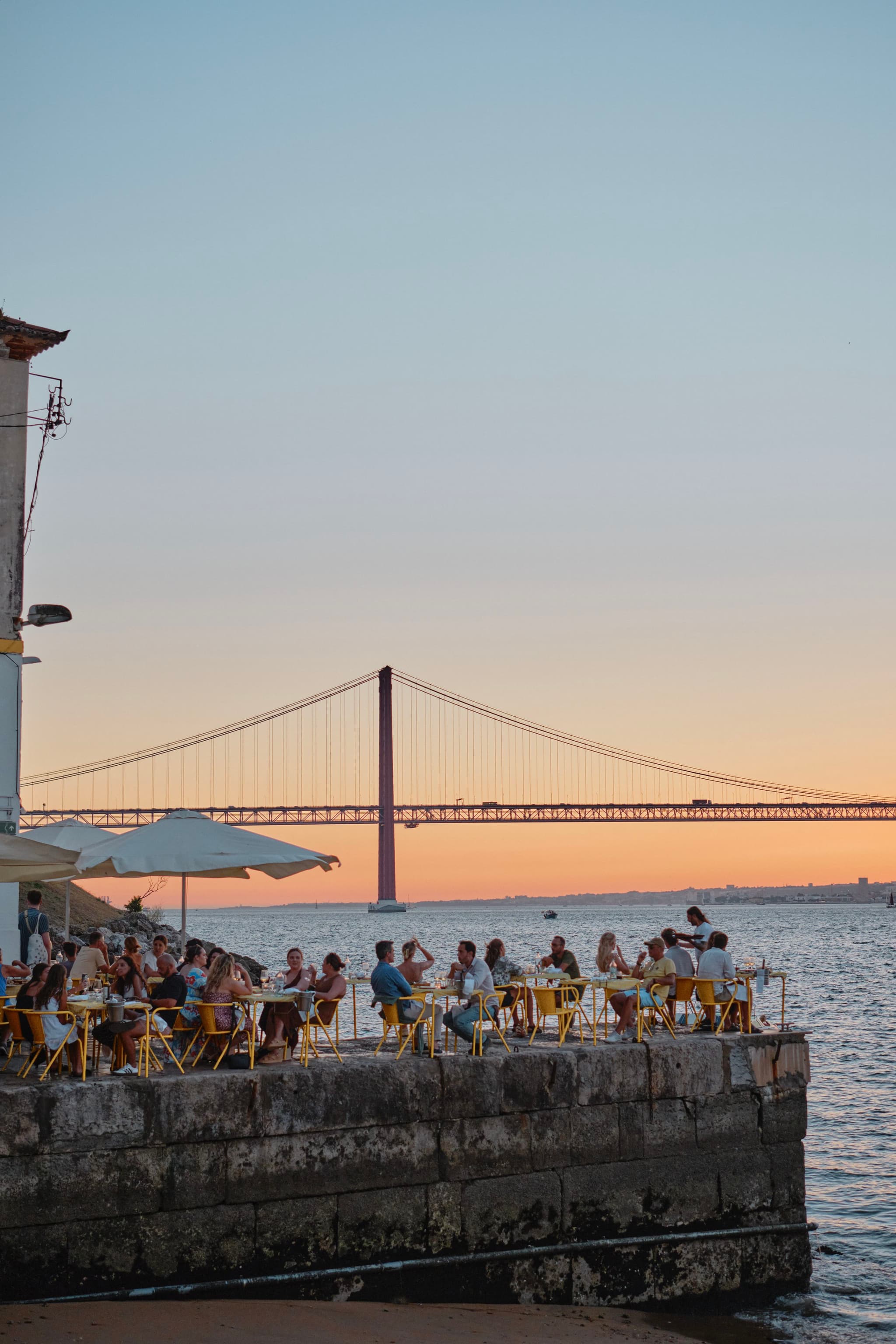 People sitting at an outdoor café by the water, with a large suspension bridge in the background during sunset