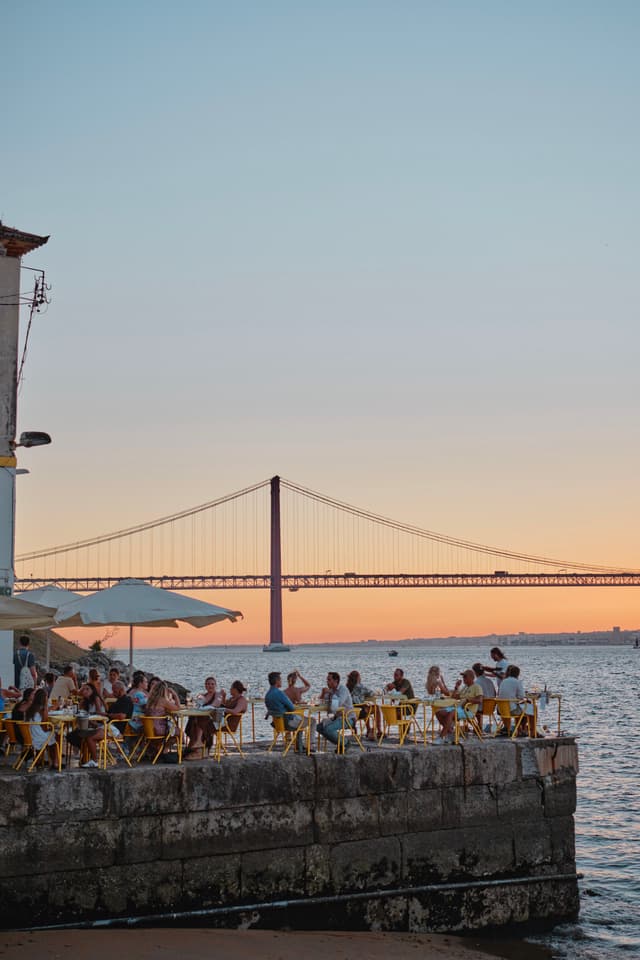 People sitting at an outdoor café by the water, with a large suspension bridge in the background during sunset