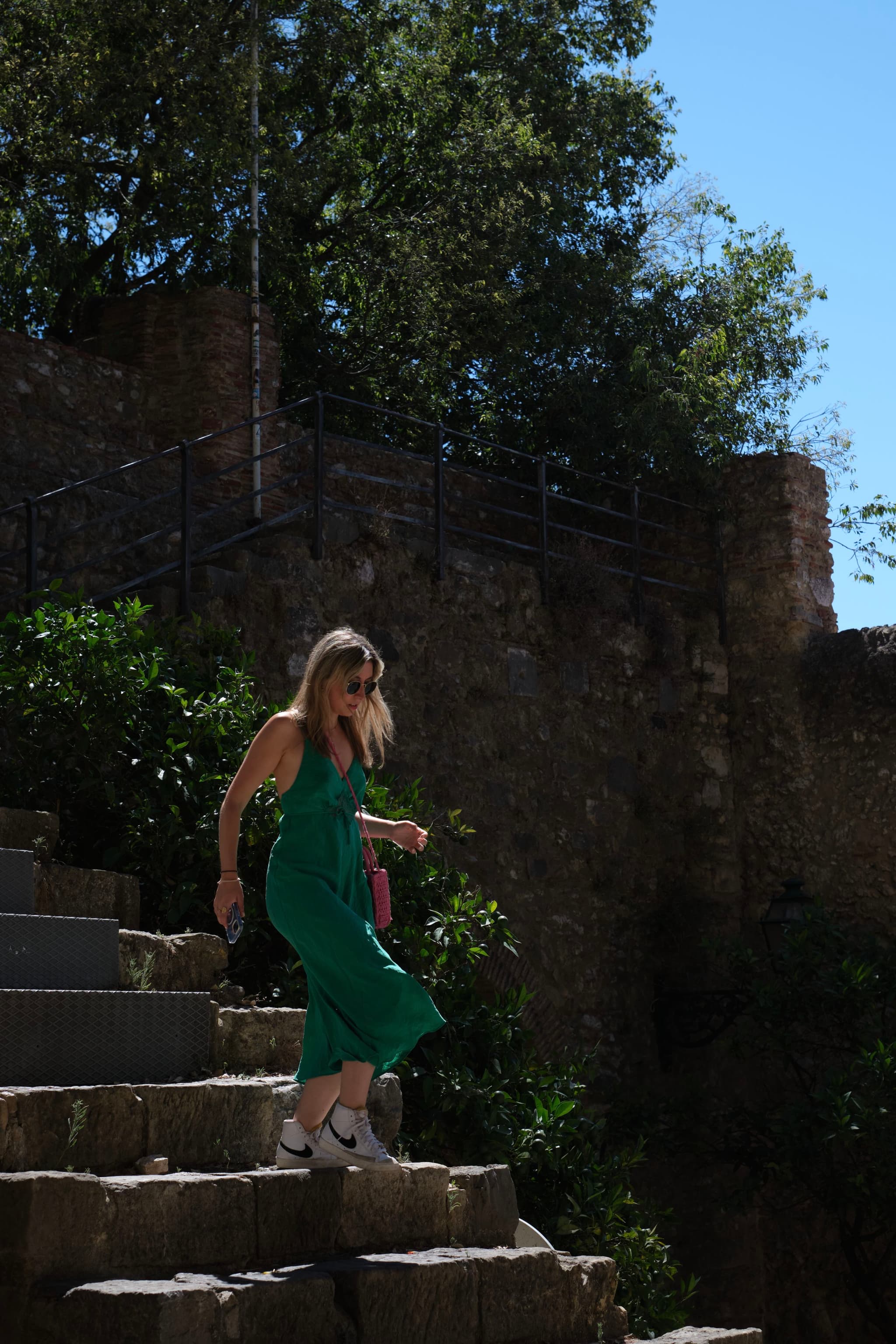A woman in a green dress walks down stone steps surrounded by greenery and a stone wall under a clear blue sky
