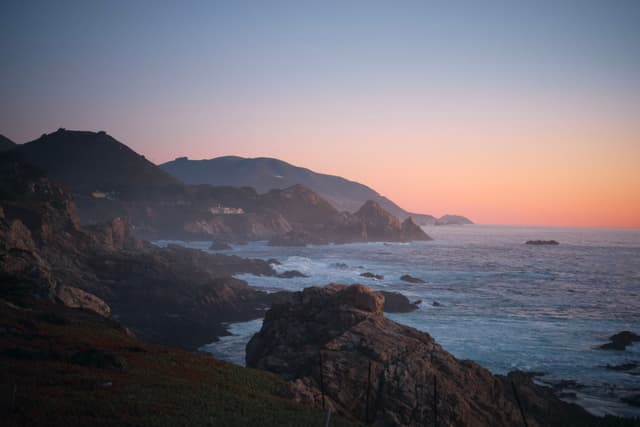 A rocky coastline at sunset with waves crashing against the cliffs and a gradient sky transitioning from orange to blue