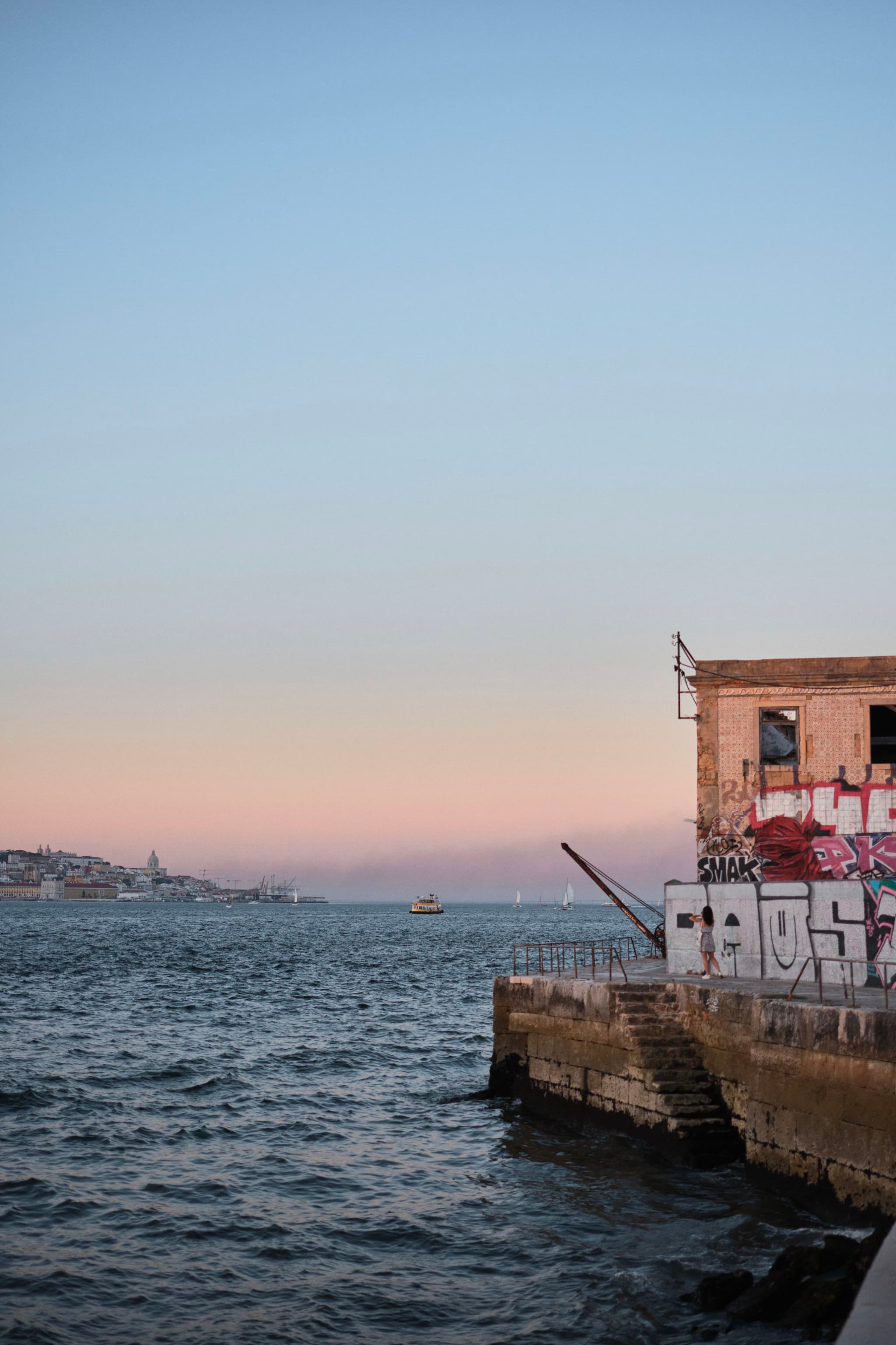 A waterfront scene at sunset with a graffiti-covered building on the right, overlooking a body of water. A distant cityscape is visible on the horizon under a clear sky