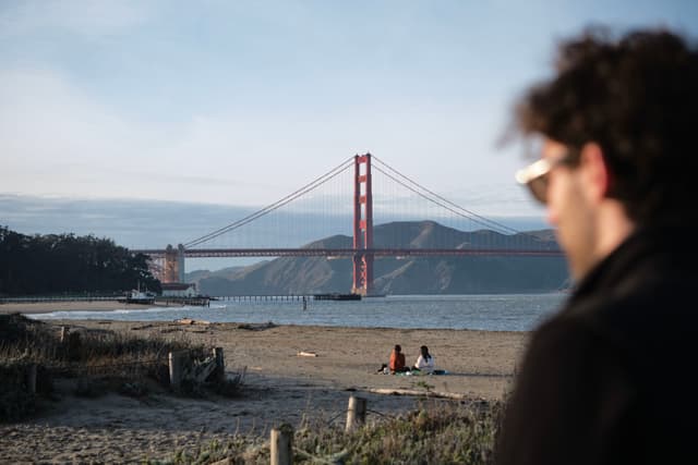 A person in sunglasses is in the foreground with the Golden Gate Bridge in the background, viewed from a beach with a few people sitting on the sand