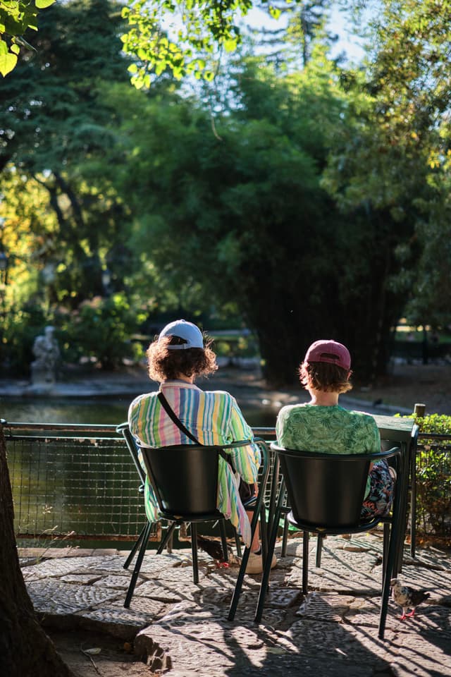 Two people sitting on chairs by a pond in a lush, green park, with sunlight filtering through the trees