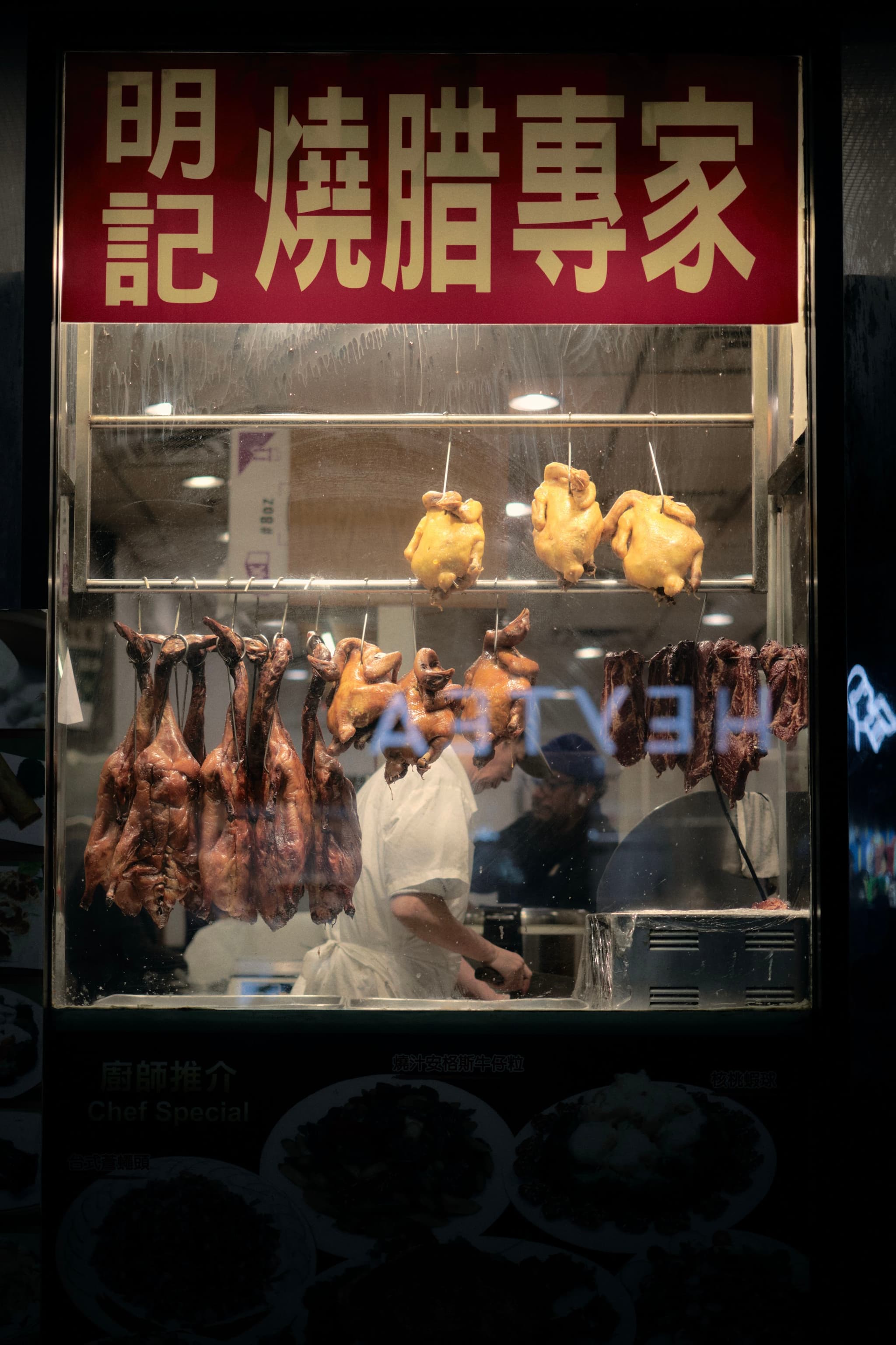 A display window with hanging roasted meats, including ducks and chickens, and a person working inside. A sign with Chinese characters is above the window