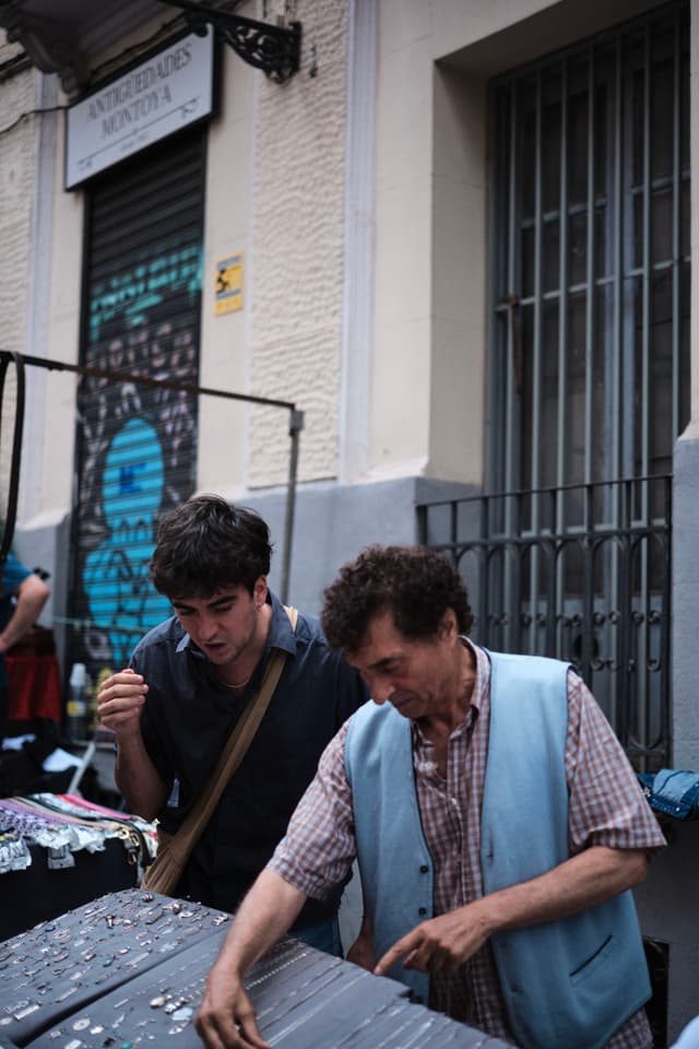 Two people are examining items on a street market table, with a building and graffiti in the background