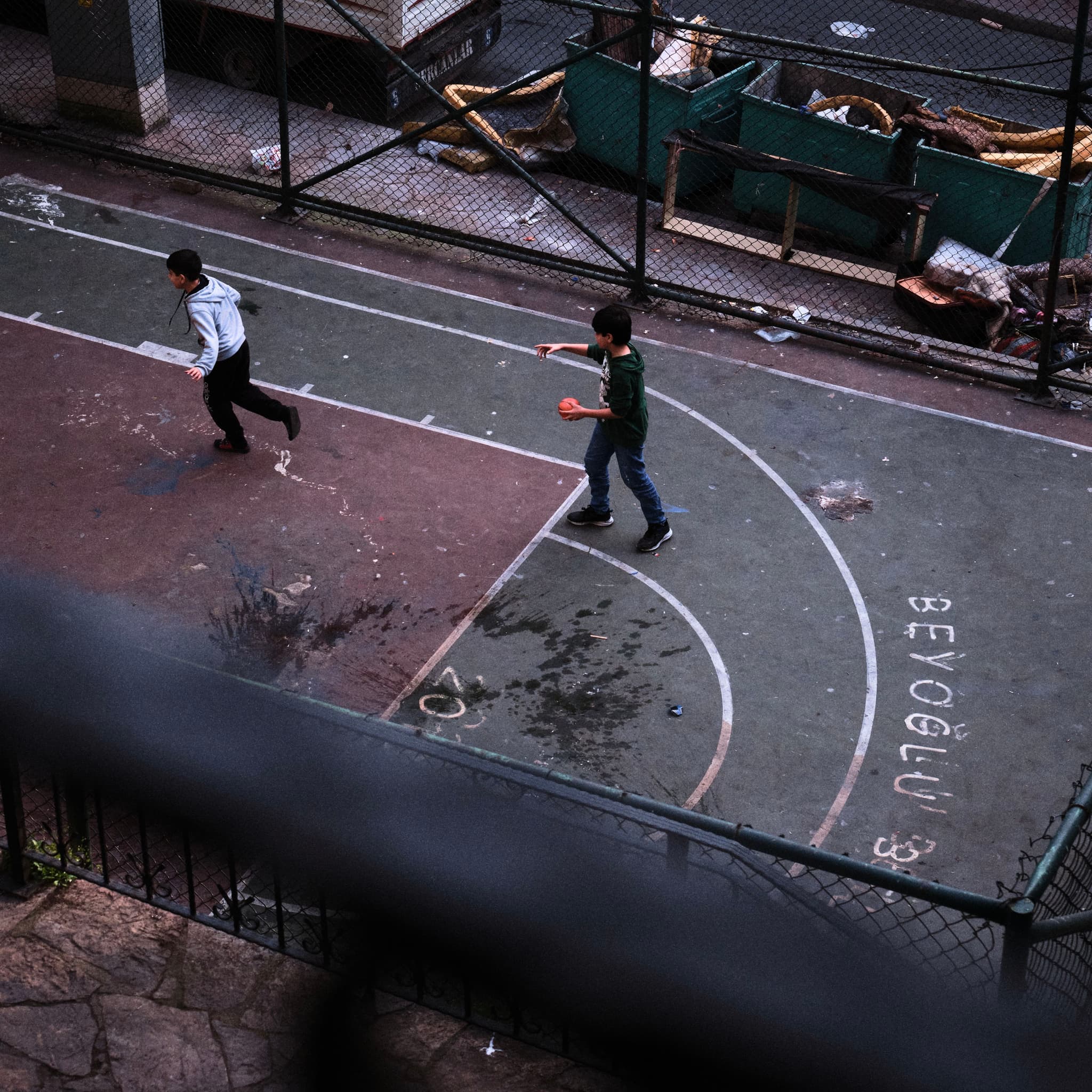 Two people running on a wet, outdoor basketball court with a fence and some debris in the background