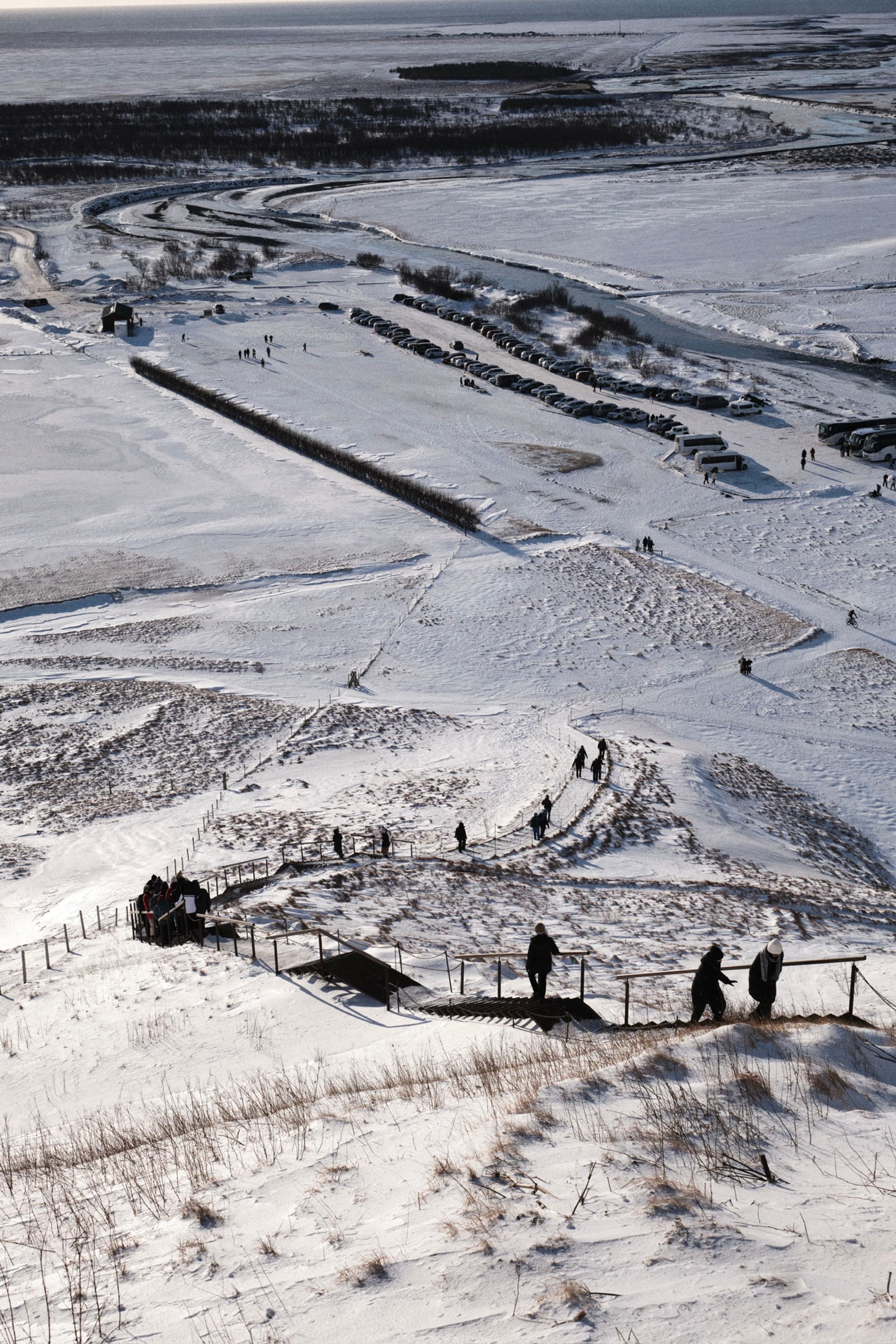 A snowy landscape with people walking along a path, featuring a series of steps and railings. The background includes a frozen body of water and distant land formations