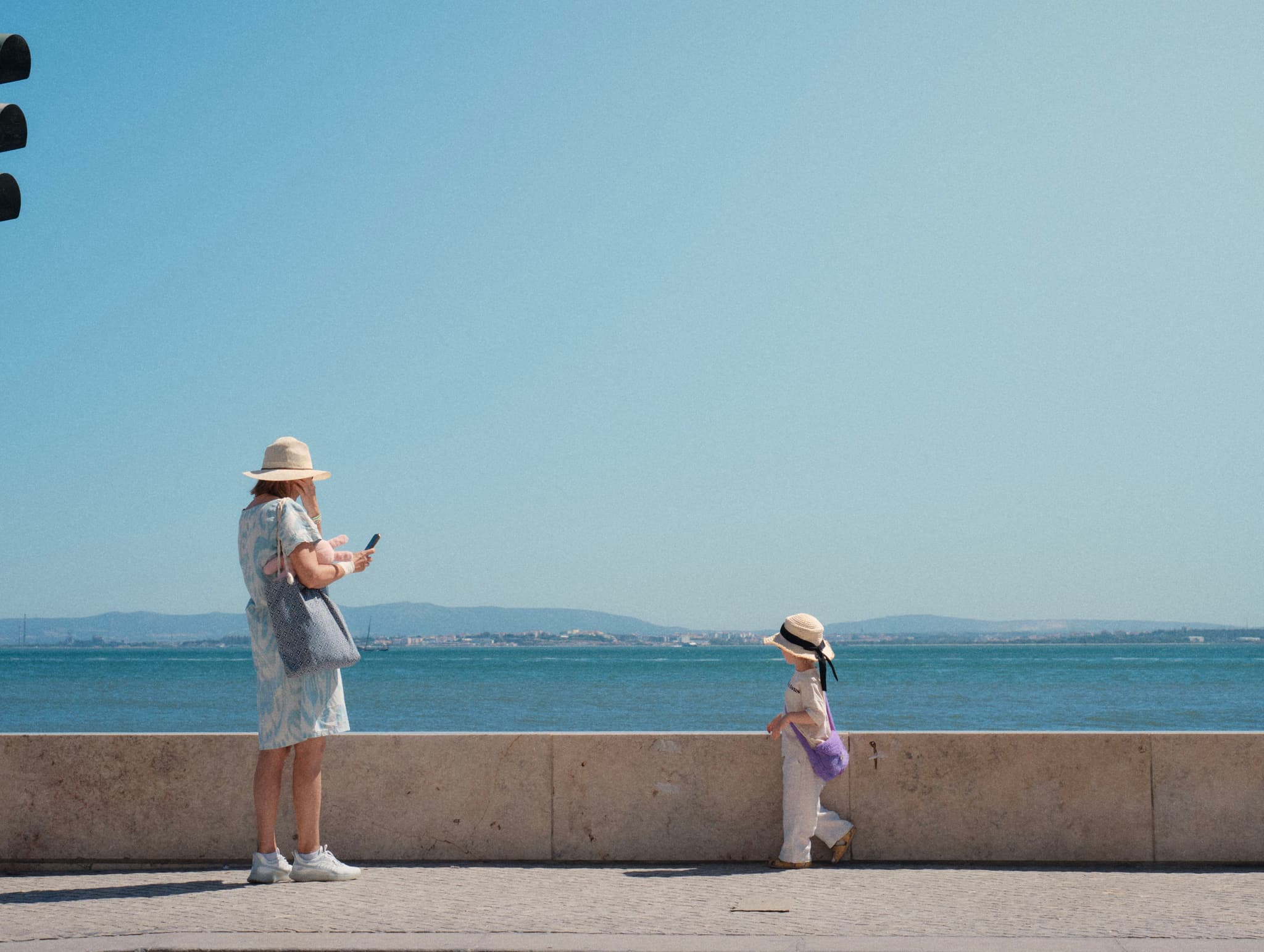 A woman and a child wearing hats stand by a waterfront, with the woman looking at her phone and the child gazing at the water. The background features a clear blue sky and distant hills
