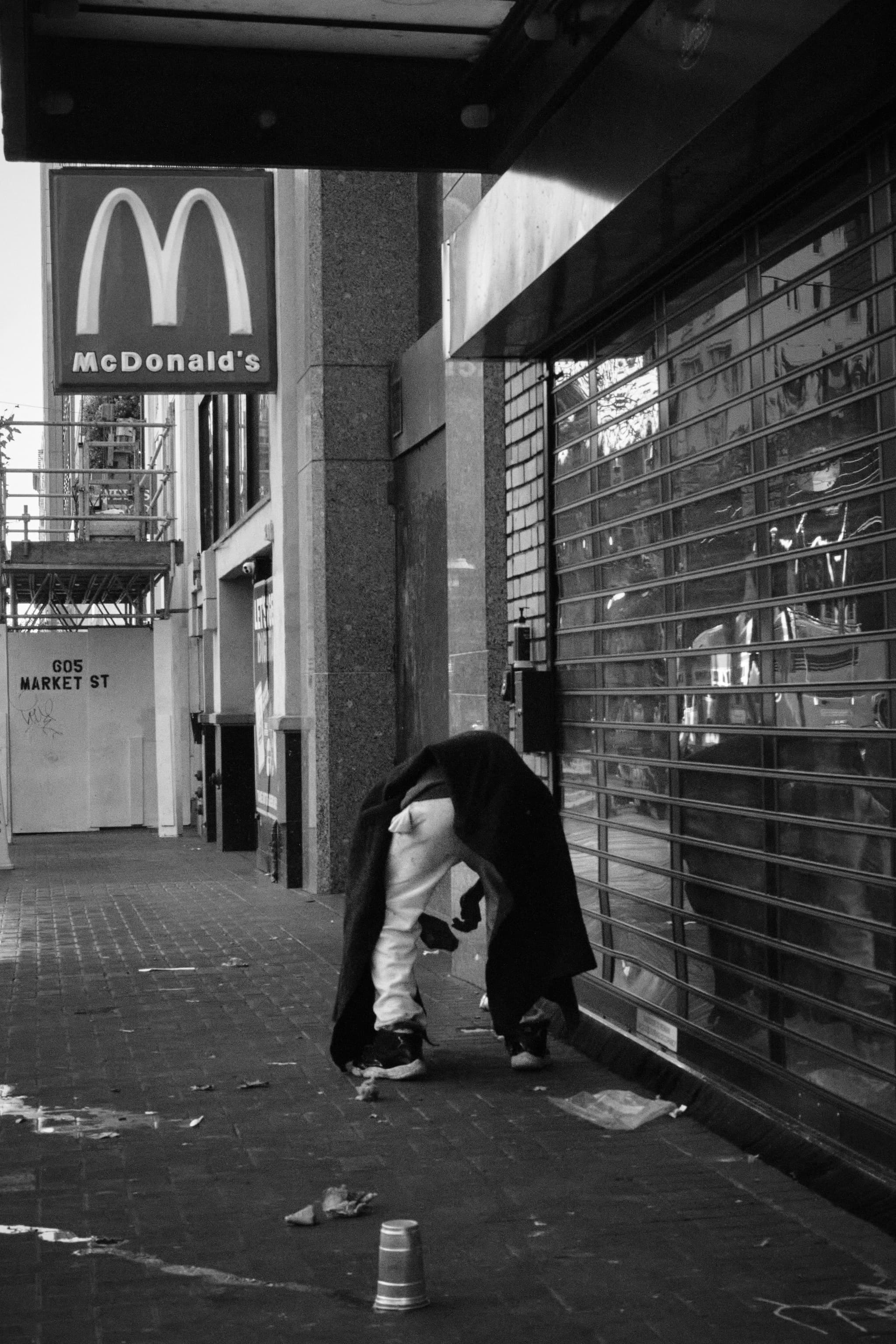 A person bends over on a city sidewalk near a closed storefront with a metal shutter. A McDonald's sign is visible in the background, and there are scattered items on the ground