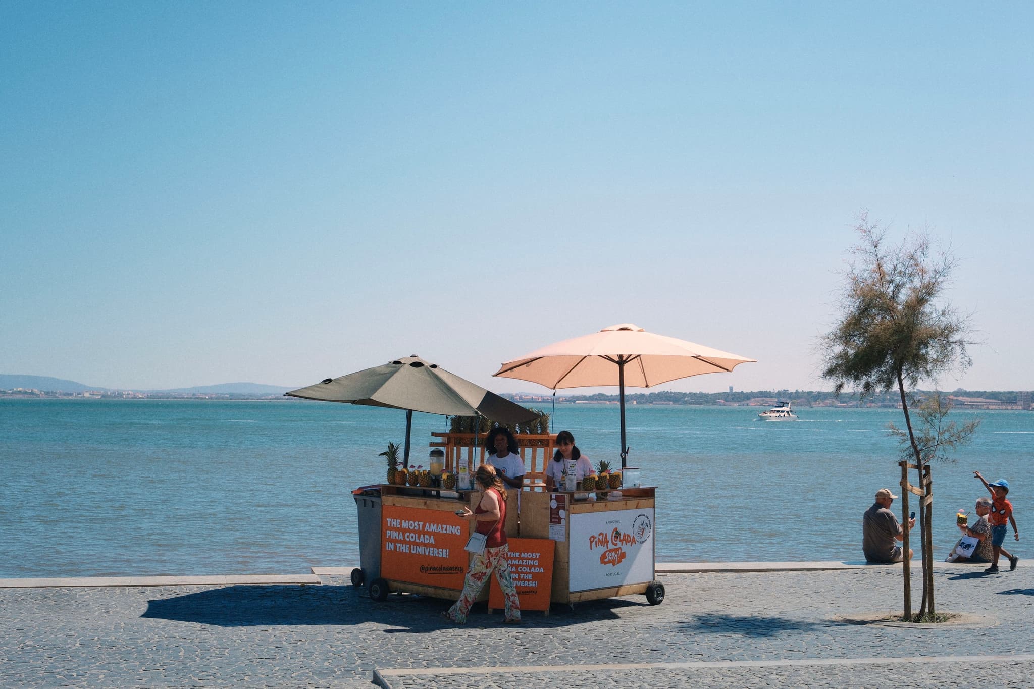 A small food cart with umbrellas is set up by a waterfront, with a few people nearby and a clear blue sky overhead