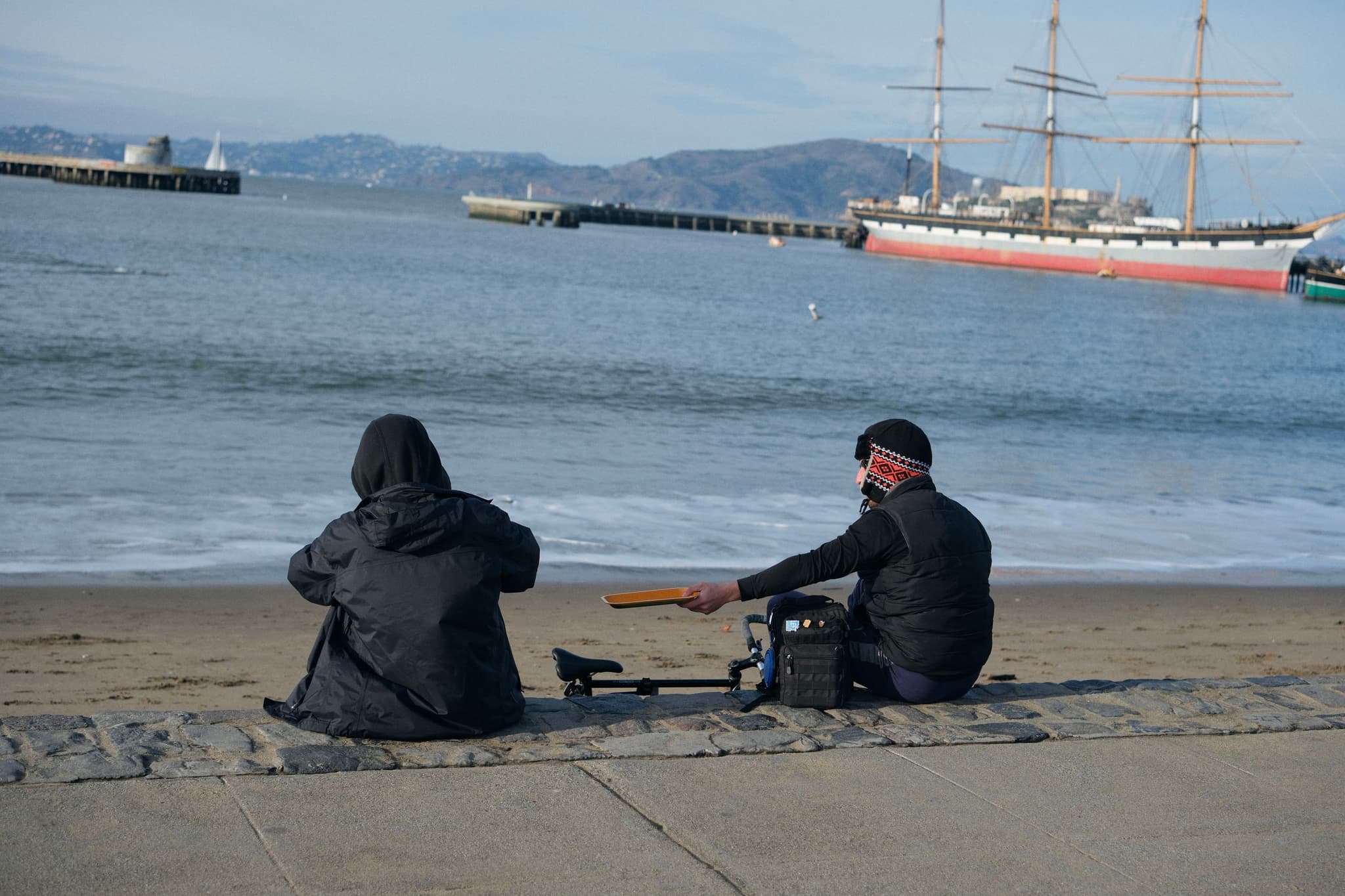 Two people sitting on a beach facing the ocean, with a large sailing ship in the background. One person is holding a guitar