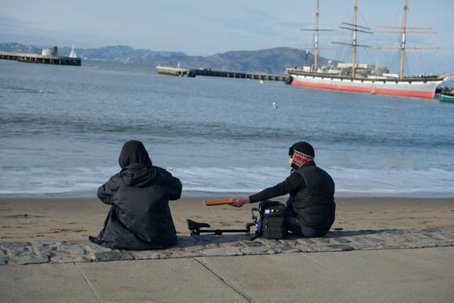 Two people sitting on a beach facing the ocean, with a large sailing ship in the background. One person is holding a guitar