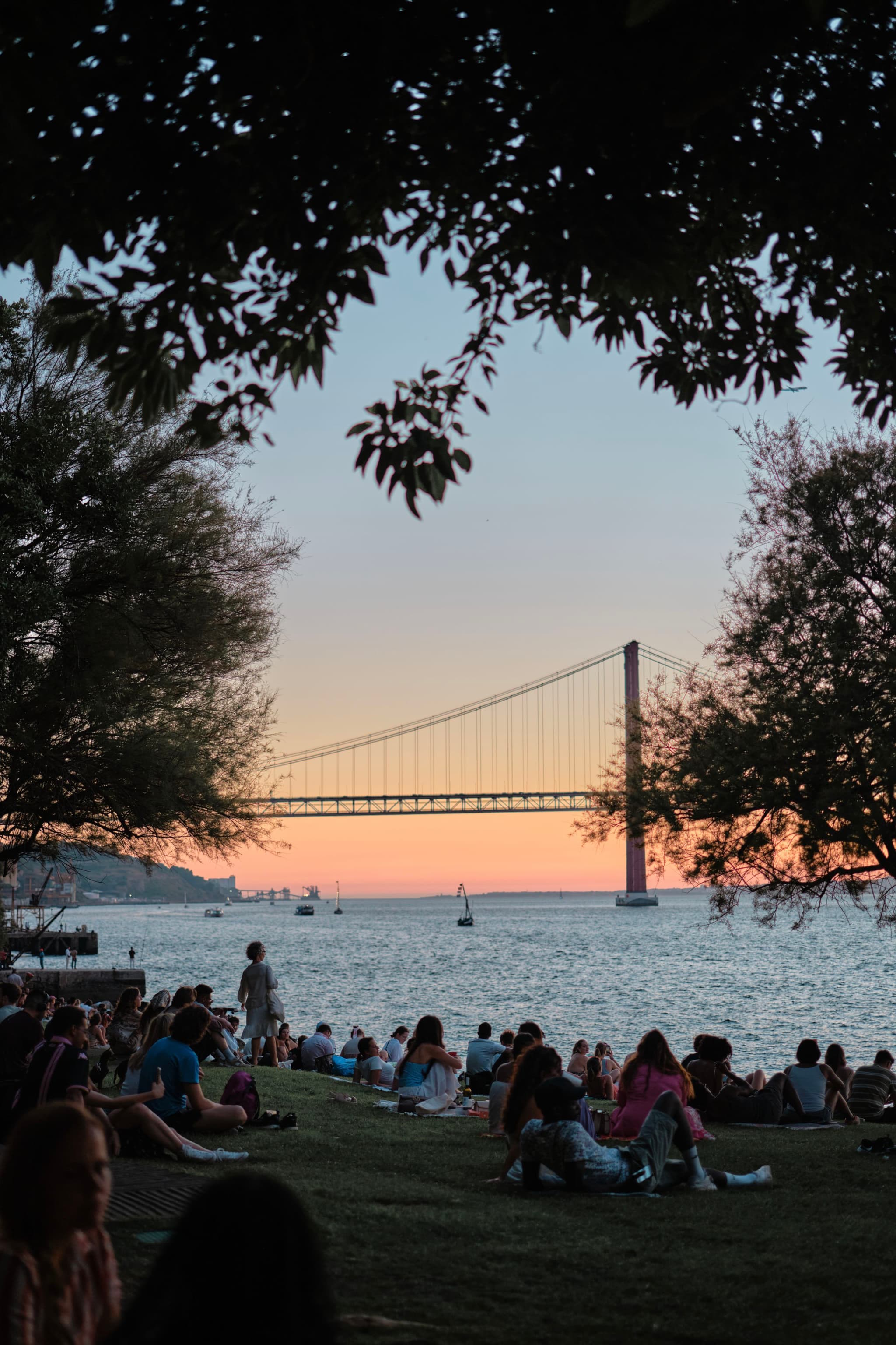 A scenic view of a bridge at sunset, with people gathered on a grassy area by the water, framed by trees
