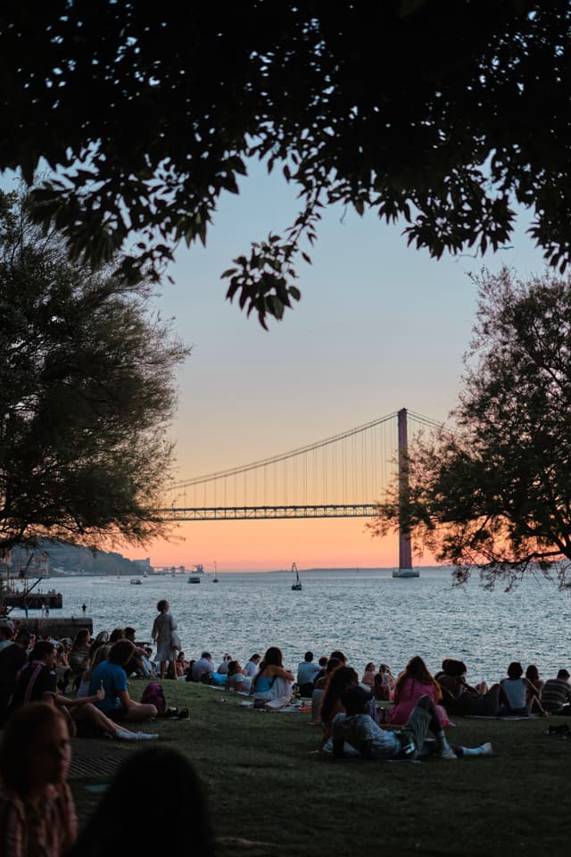A scenic view of a bridge at sunset, with people gathered on a grassy area by the water, framed by trees