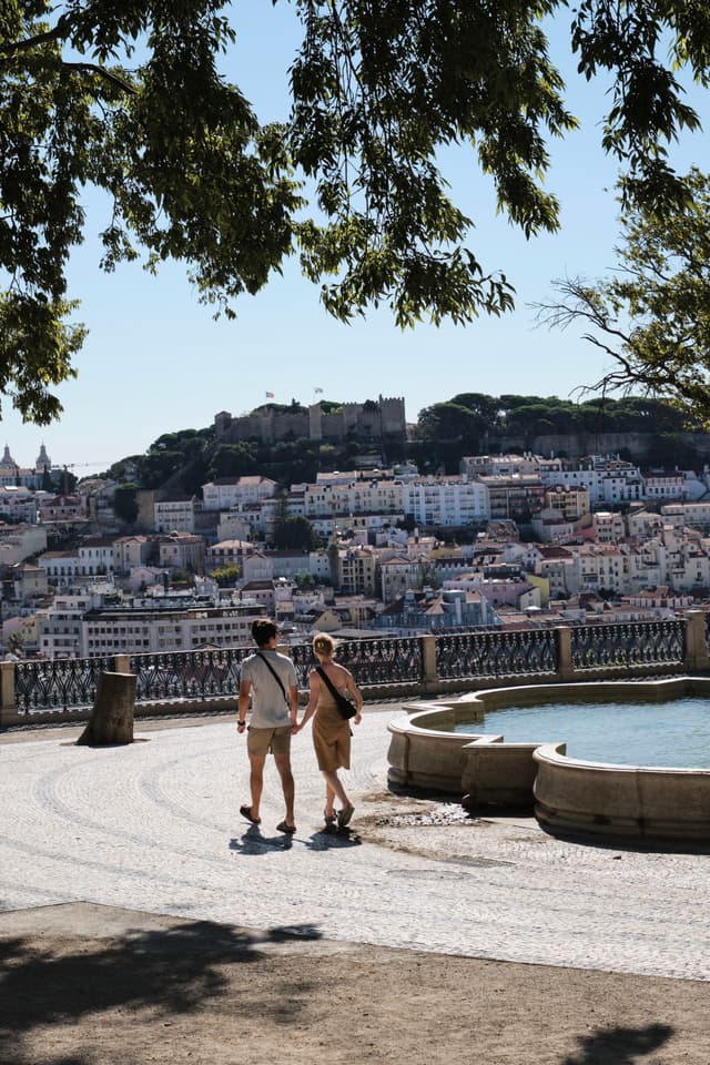 Two people walking near a circular fountain with a scenic view of a city and a hilltop castle in the background, framed by tree branches