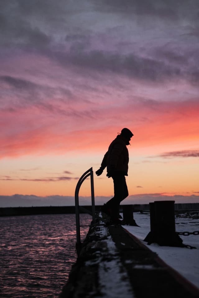 A silhouette of a person walking along a pier at sunset, with a vibrant sky of pink and purple hues reflected on the water