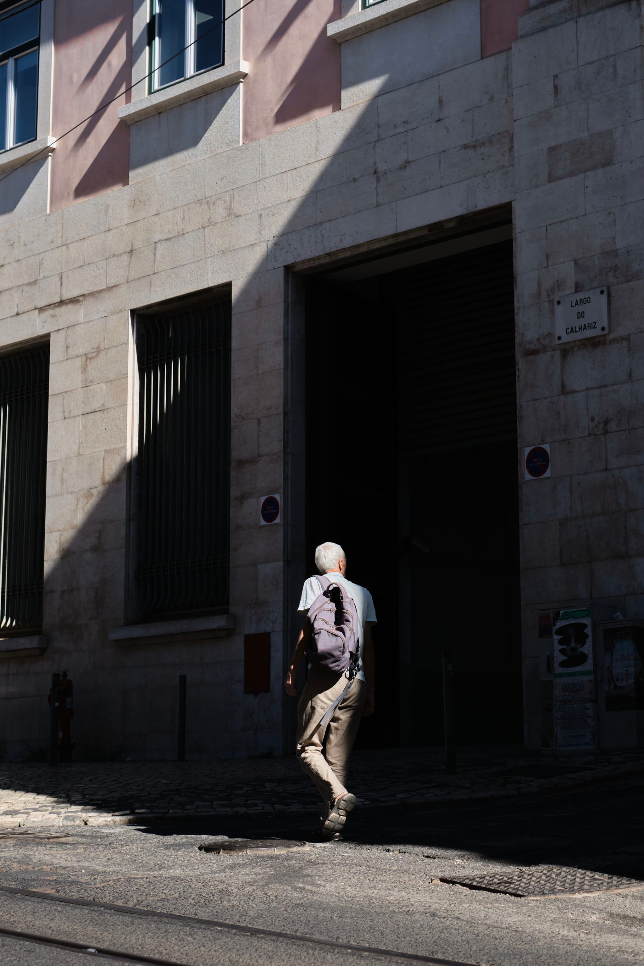 A person walking on a sunlit street, casting a shadow on a building with large windows and a dark entrance