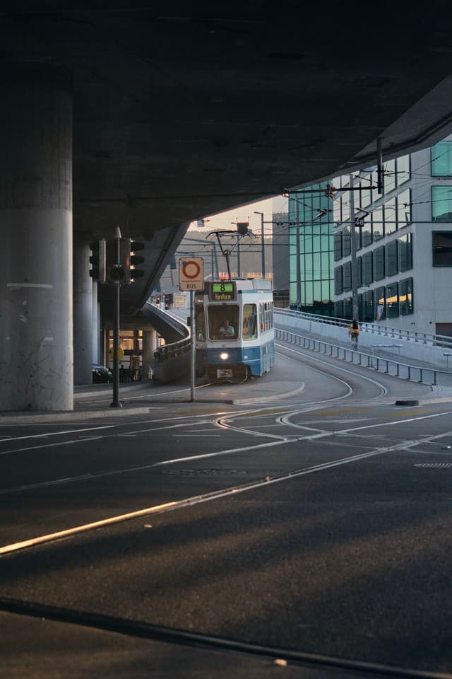 A tram navigating through an urban setting with modern buildings and an overpass, captured in low light