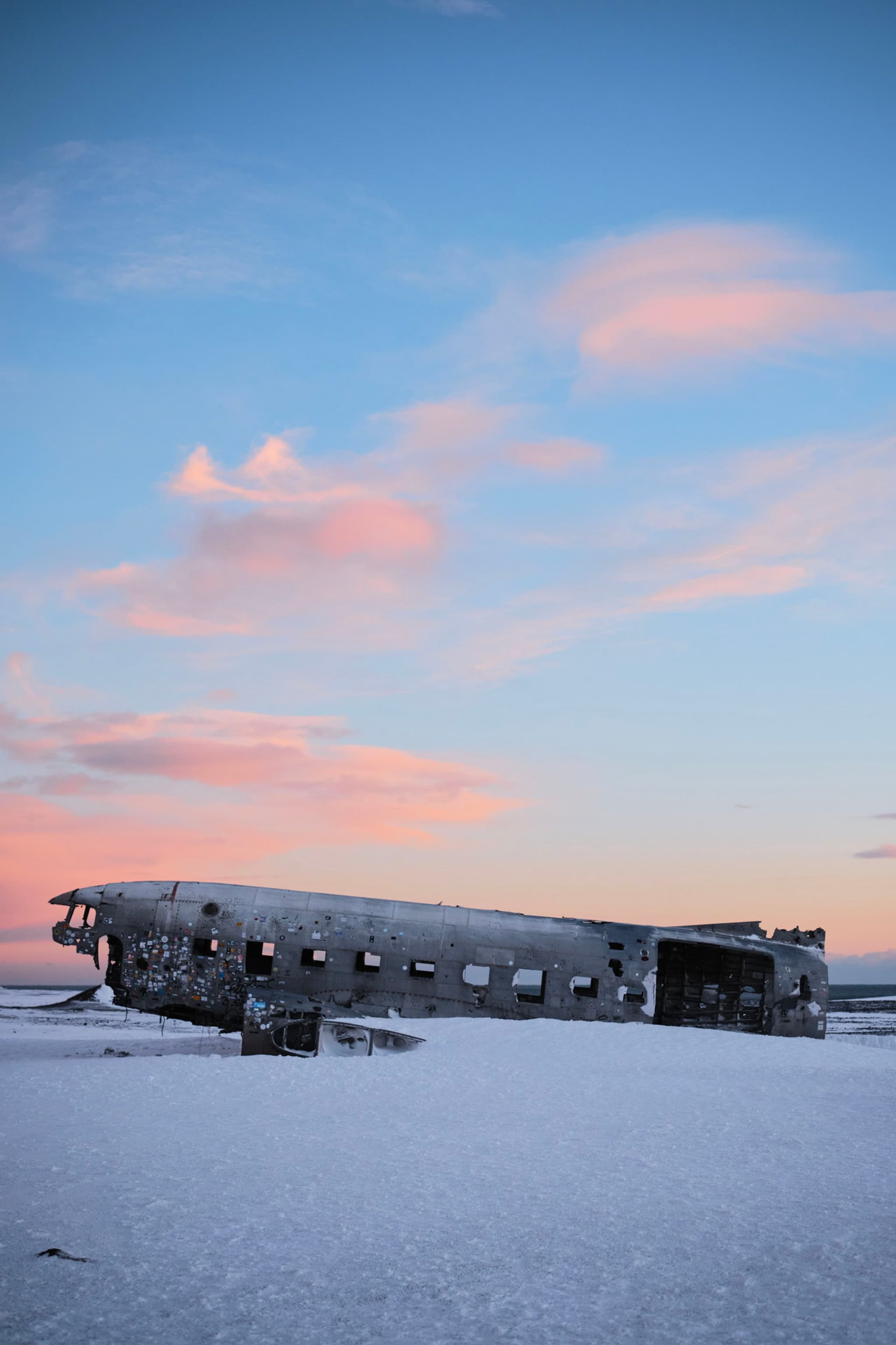 A weathered airplane wreck rests on a snowy landscape under a sky with pink-tinged clouds