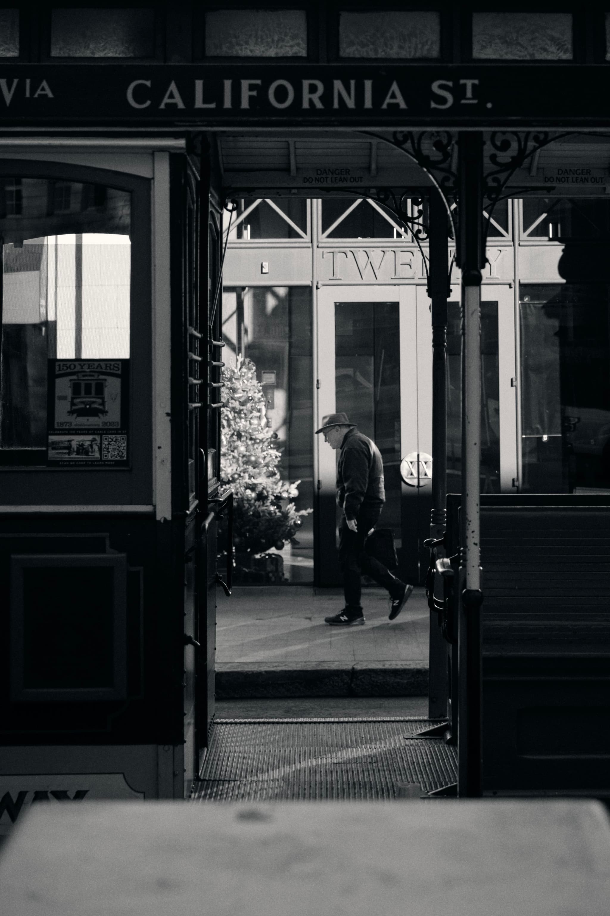 A black and white scene featuring a man walking past a shop window, viewed through the frame of a streetcar with California St. visible above