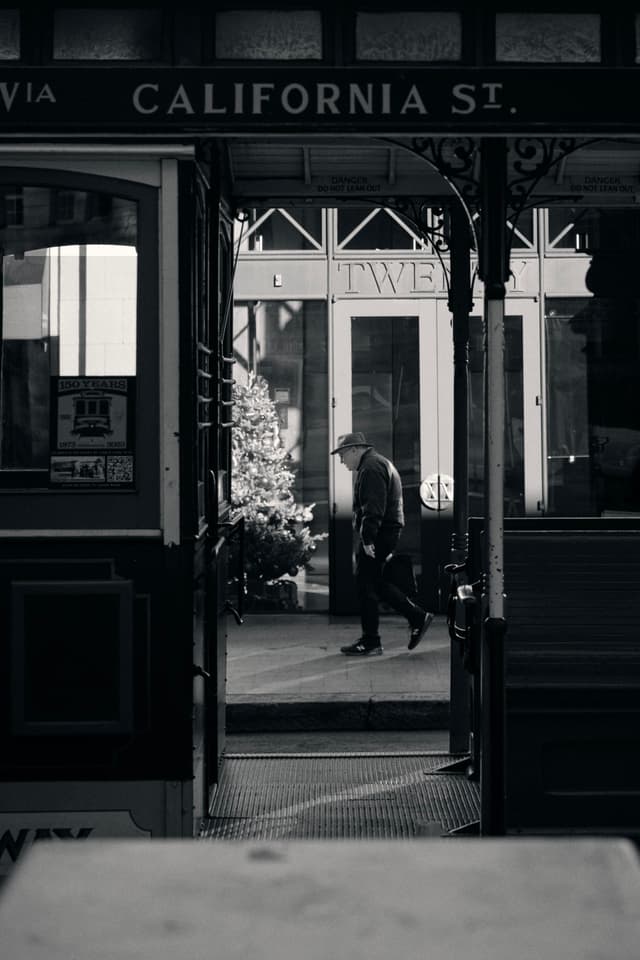 A black and white scene featuring a man walking past a shop window, viewed through the frame of a streetcar with California St. visible above